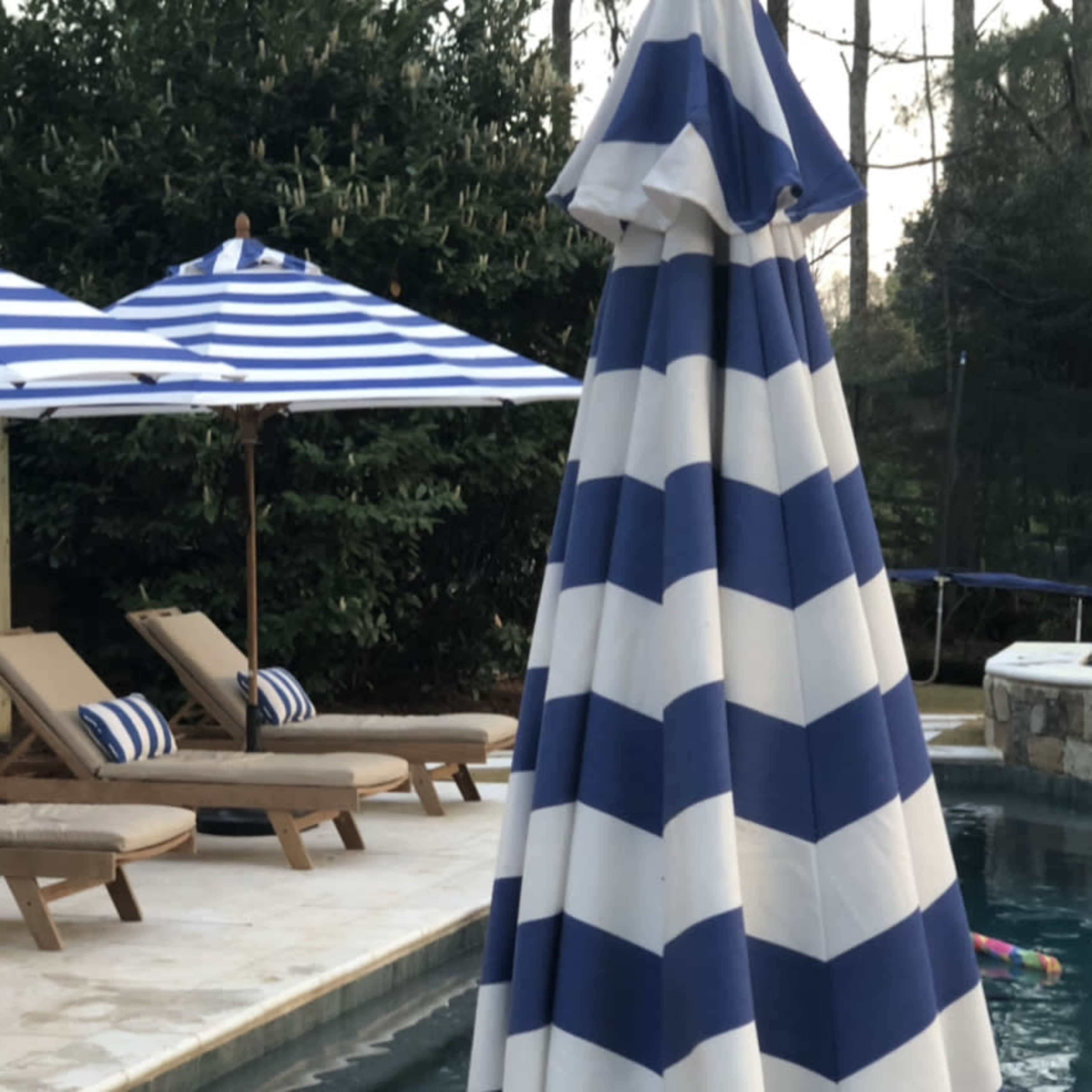 A swimming pool area with blue and white striped umbrellas, lounge chairs, and greenery in the background.