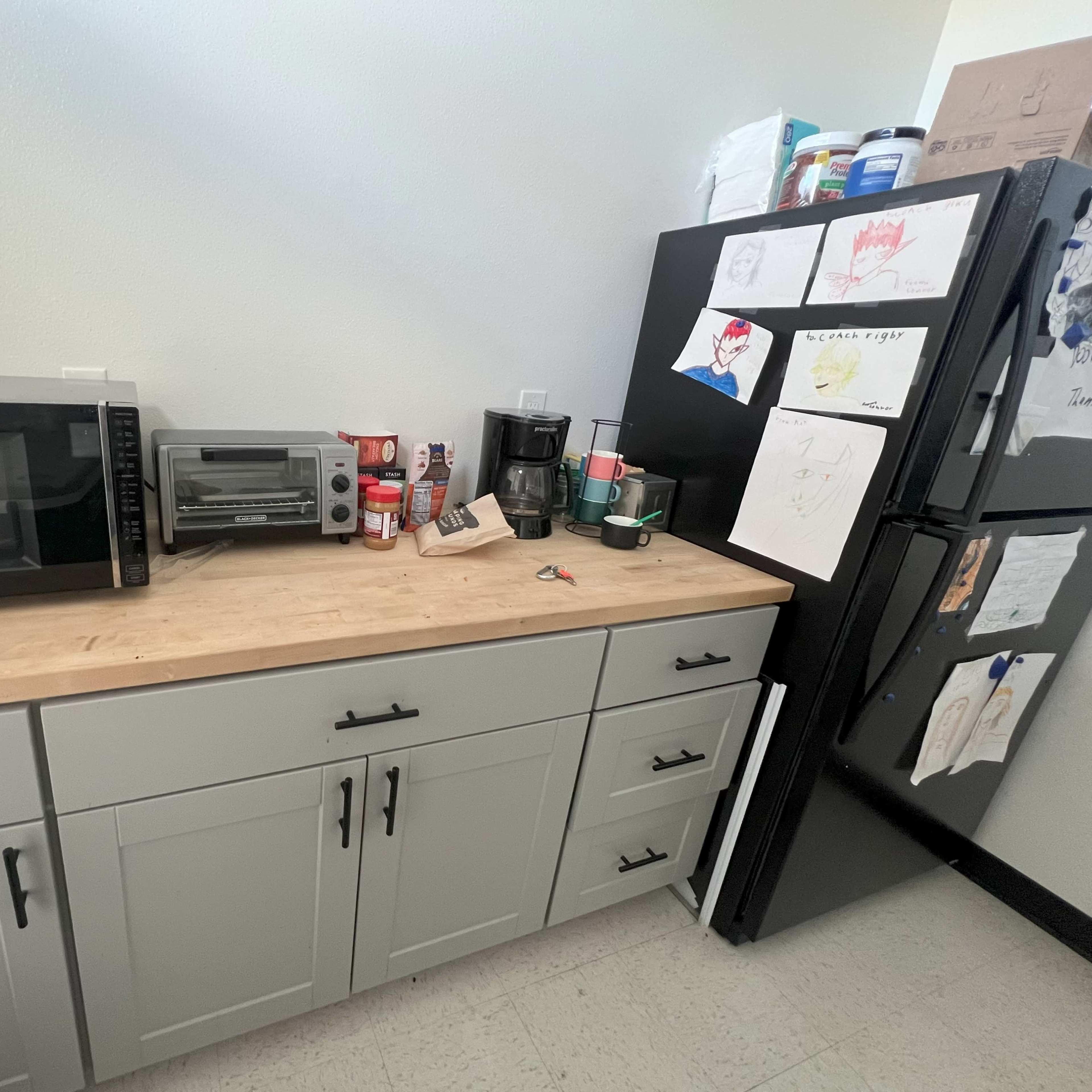 A kitchen countertop with a microwave, toaster oven, coffee maker, and various items, next to a refrigerator covered in children's drawings.