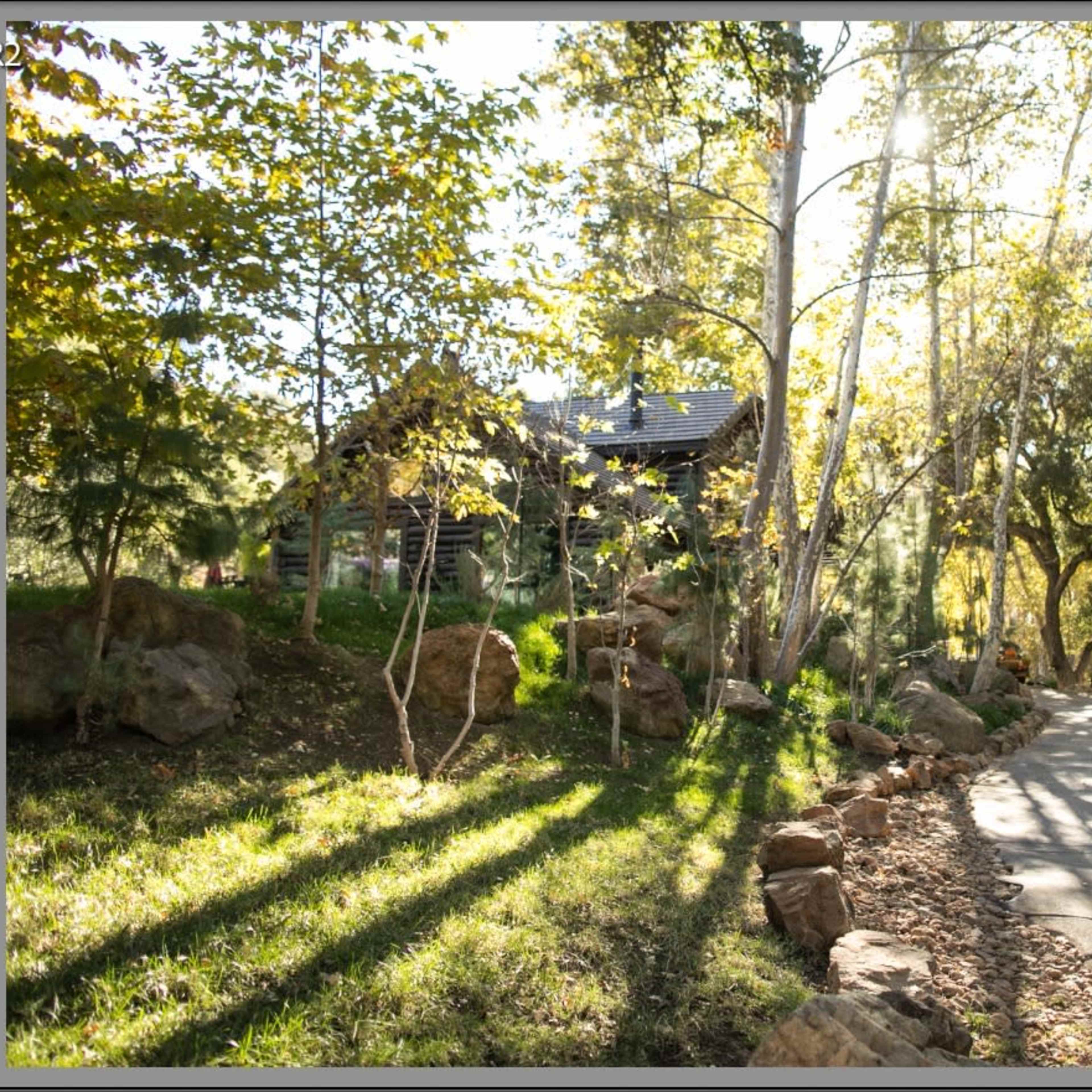 A paved pathway winds through lush greenery and rock formations, leading to a house partially obscured by trees.