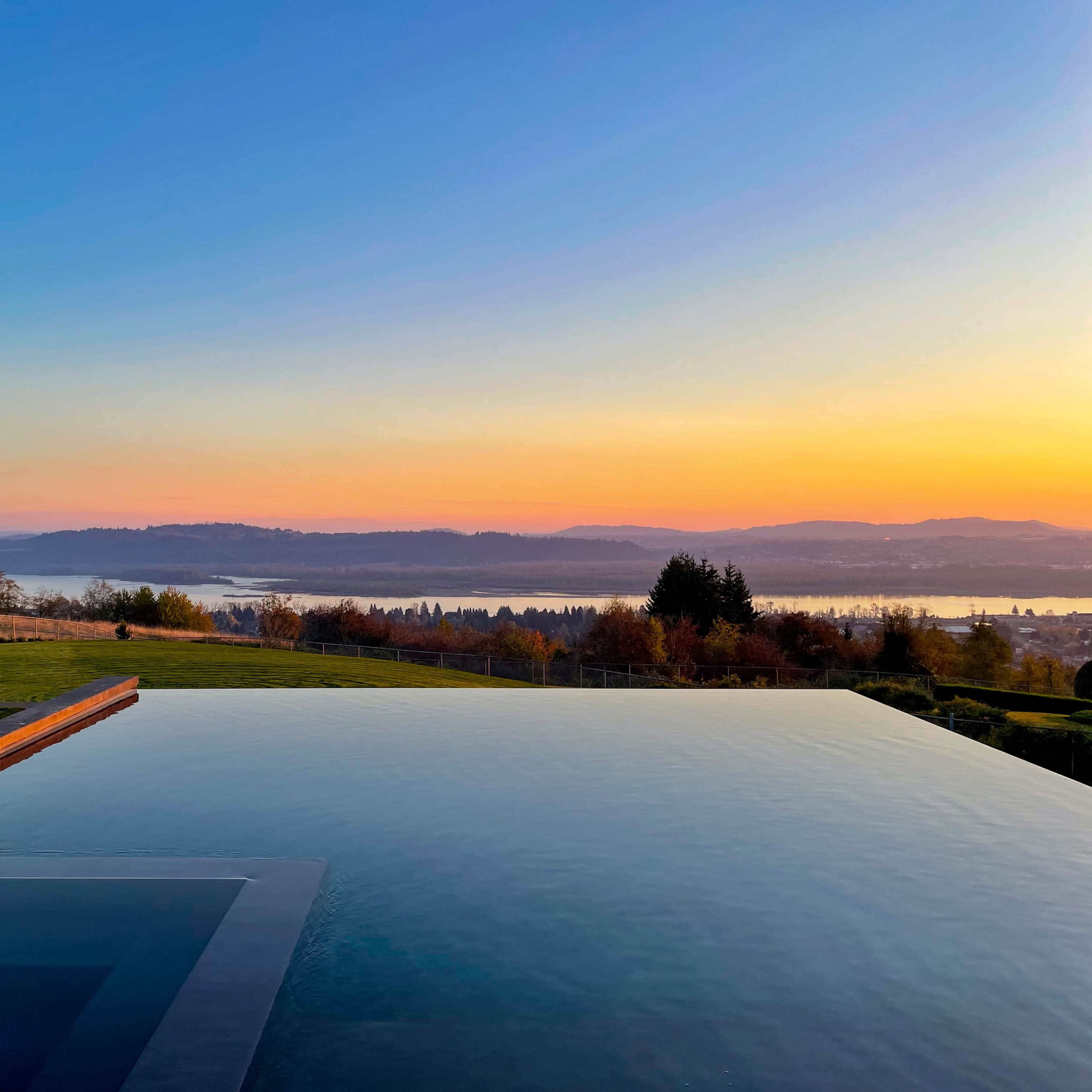 An infinity pool overlooking a landscape with rolling hills and a body of water during sunset.