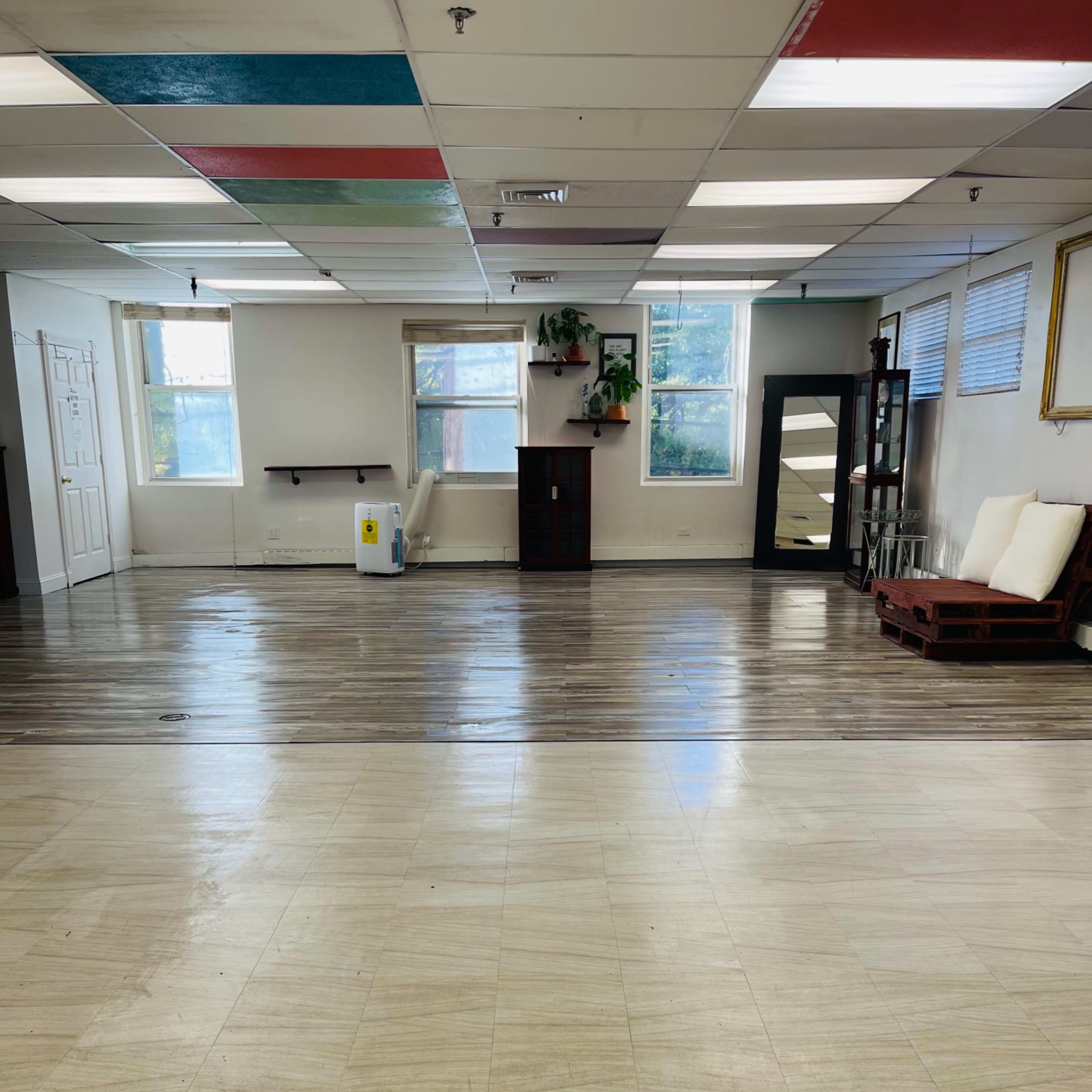 A spacious, empty room with a tiled floor, large windows, and minimal furnishings, including a mirror and a couch.