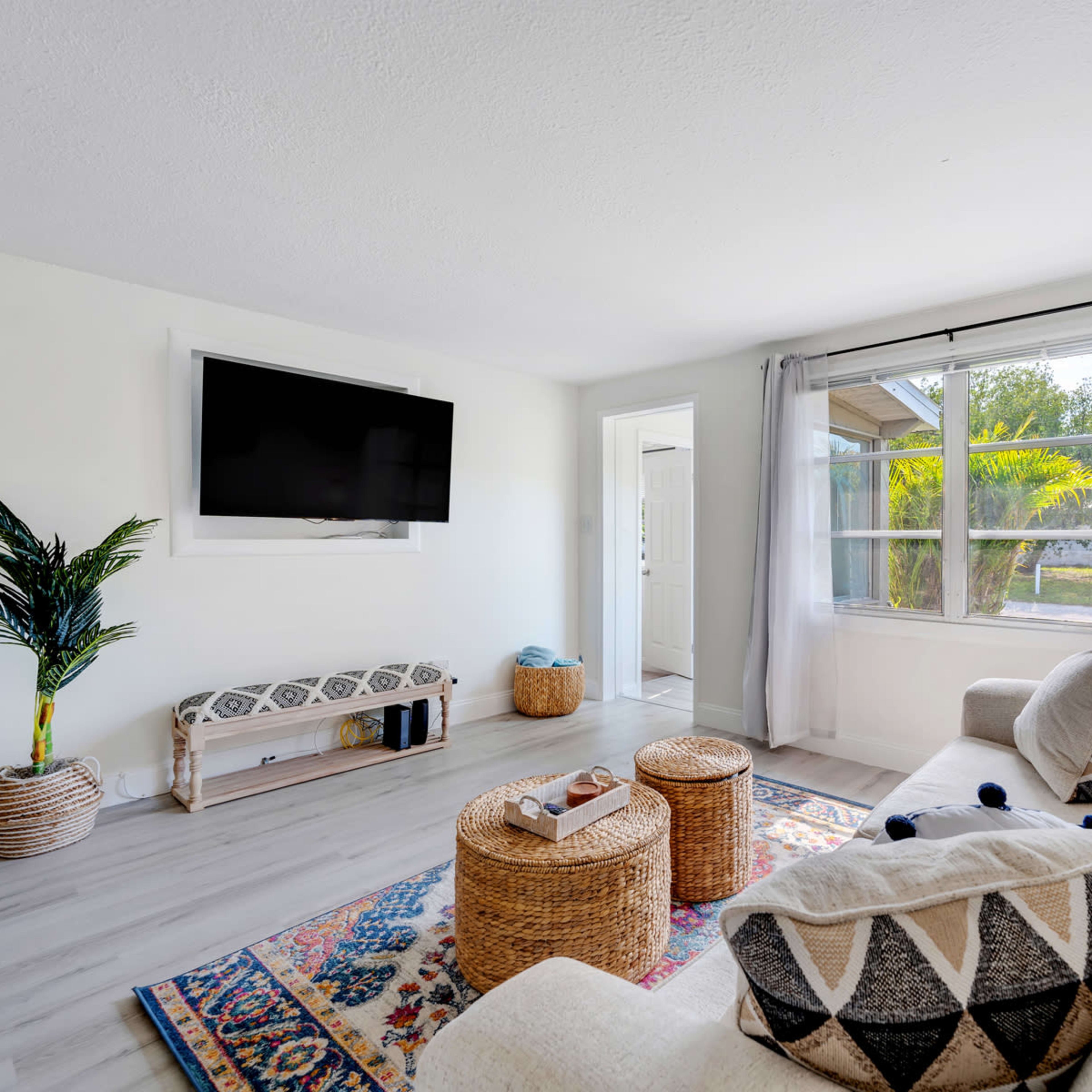 A bright living room with a large window, a comfortable sofa, a television mounted on the wall, and two round wicker tables.