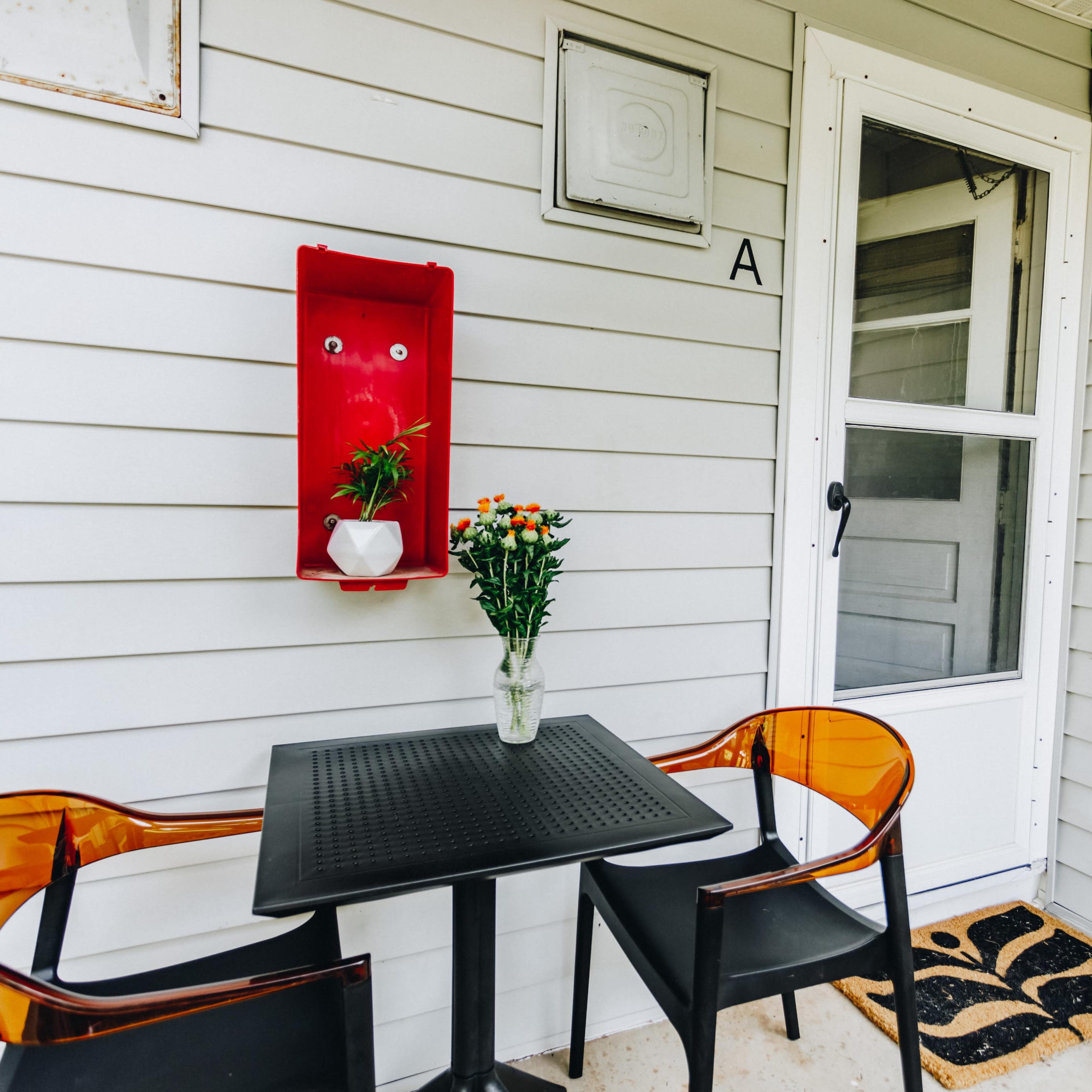 A small patio area features a black table with two transparent chairs, a flower vase, and a door leading inside.