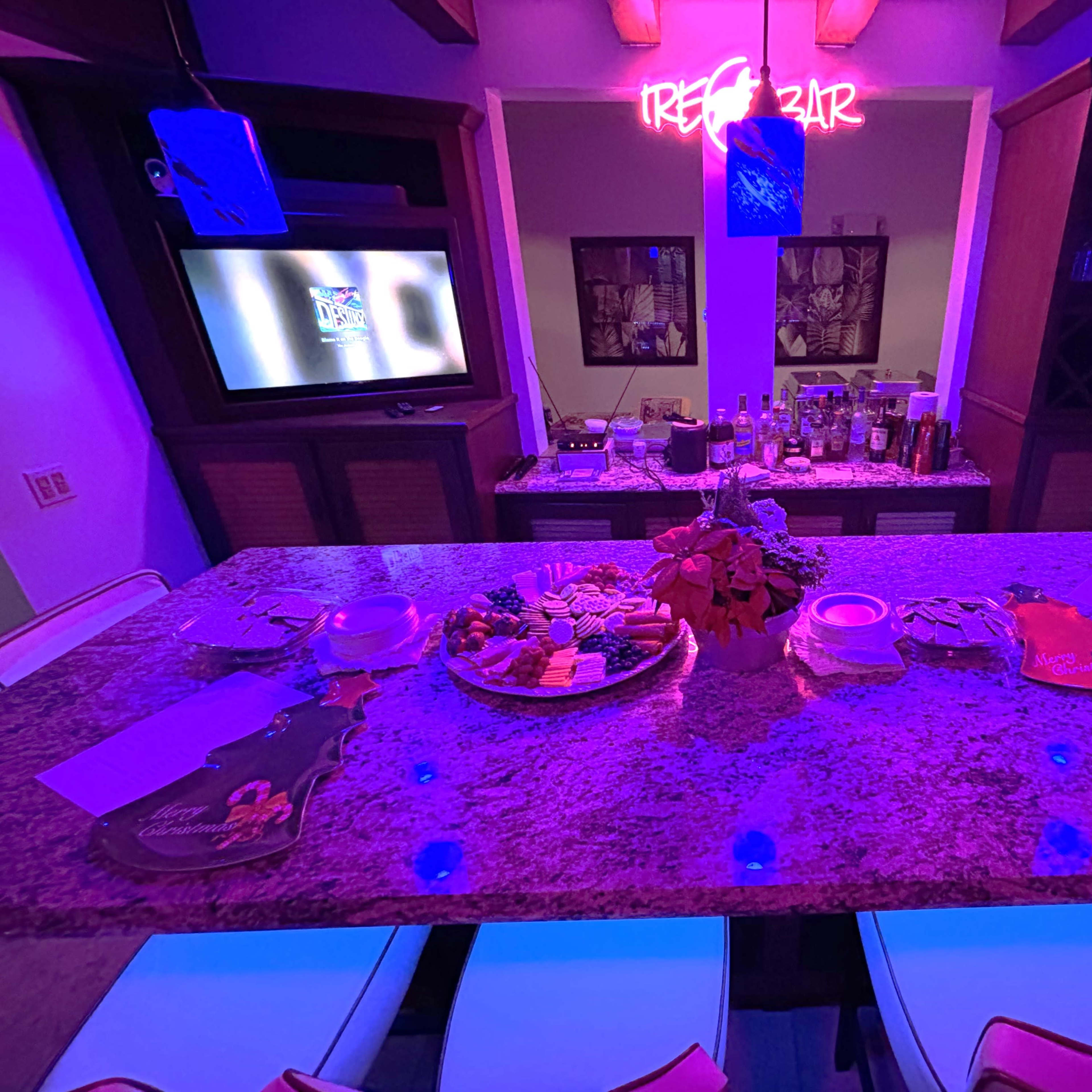 A well-lit bar area features a granite countertop displaying various appetizers and drinks, with a neon "TREZ BAR" sign in the background.