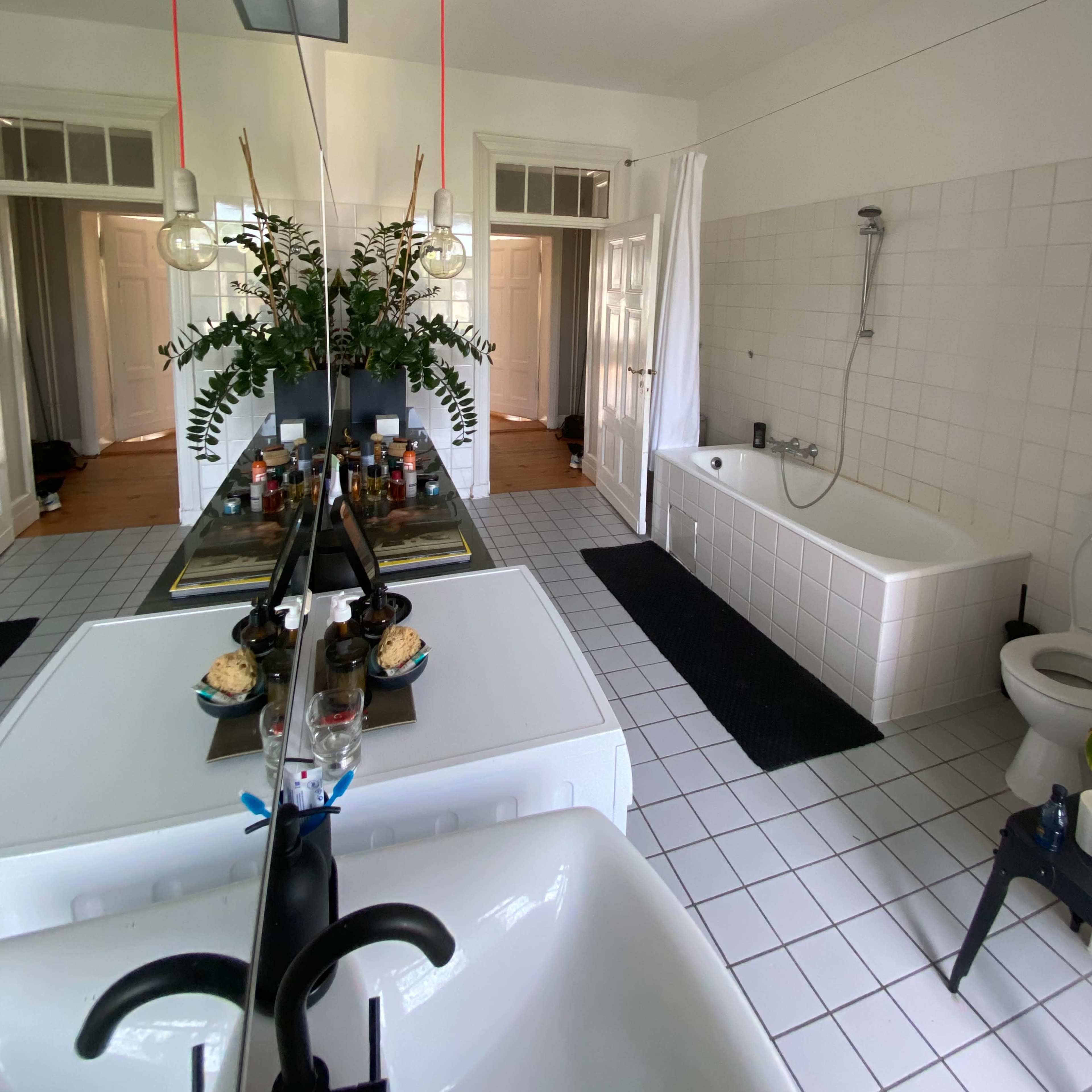A bathroom features a large mirror, a freestanding bathtub, a toilet, and a sink, with decorative plants on the counter and white tiled flooring.