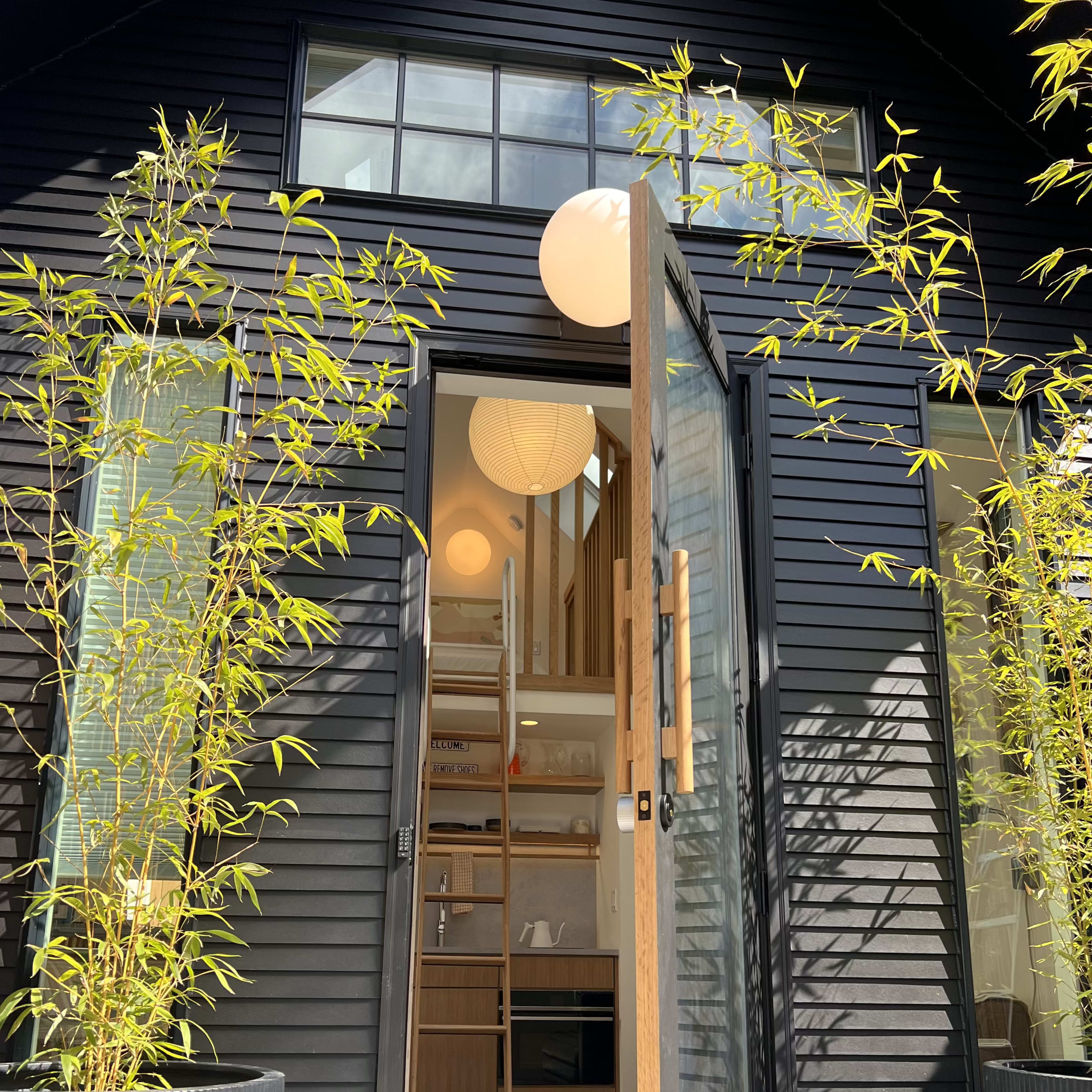 A modern black house features an open front door flanked by tall bamboo plants in planters, leading into a bright interior with a visible ladder and round pendant lights.