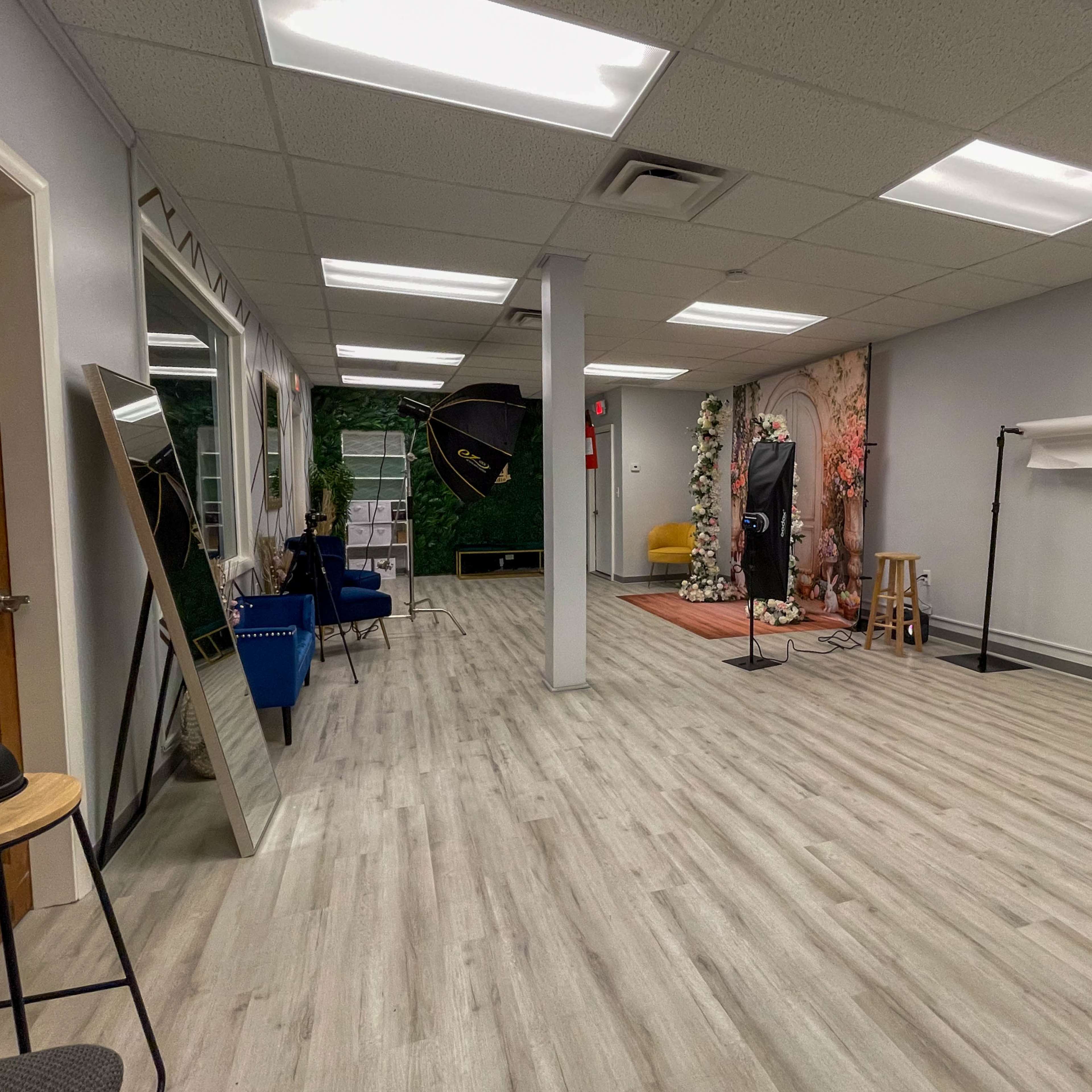 A spacious, well-lit interior of a studio with wooden flooring, featuring a photography backdrop, seating area, and various equipment set up.