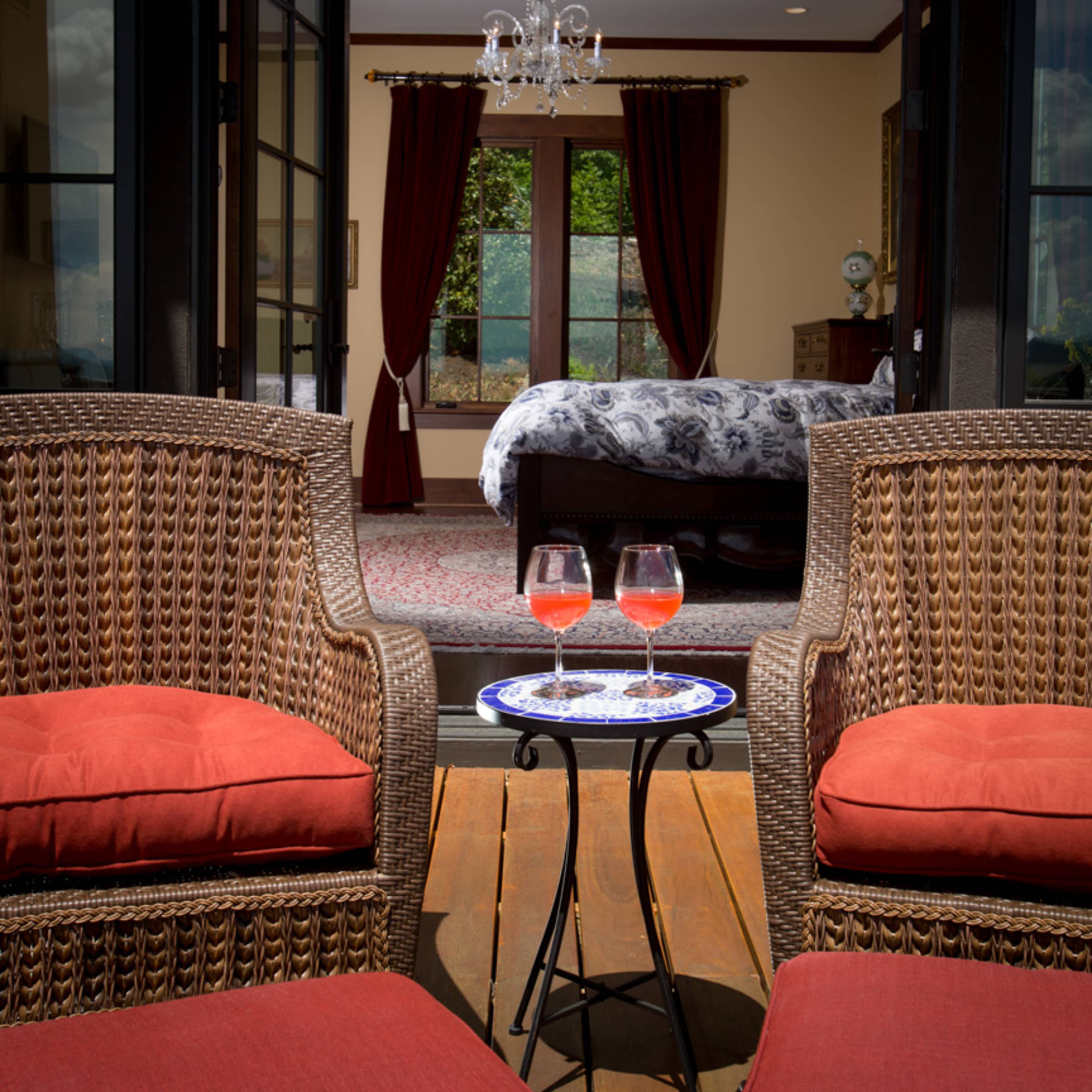 Two wicker chairs with red cushions face a small table holding two glasses of cocktail, overlooking a elegantly decorated room with a bed and large windows.
