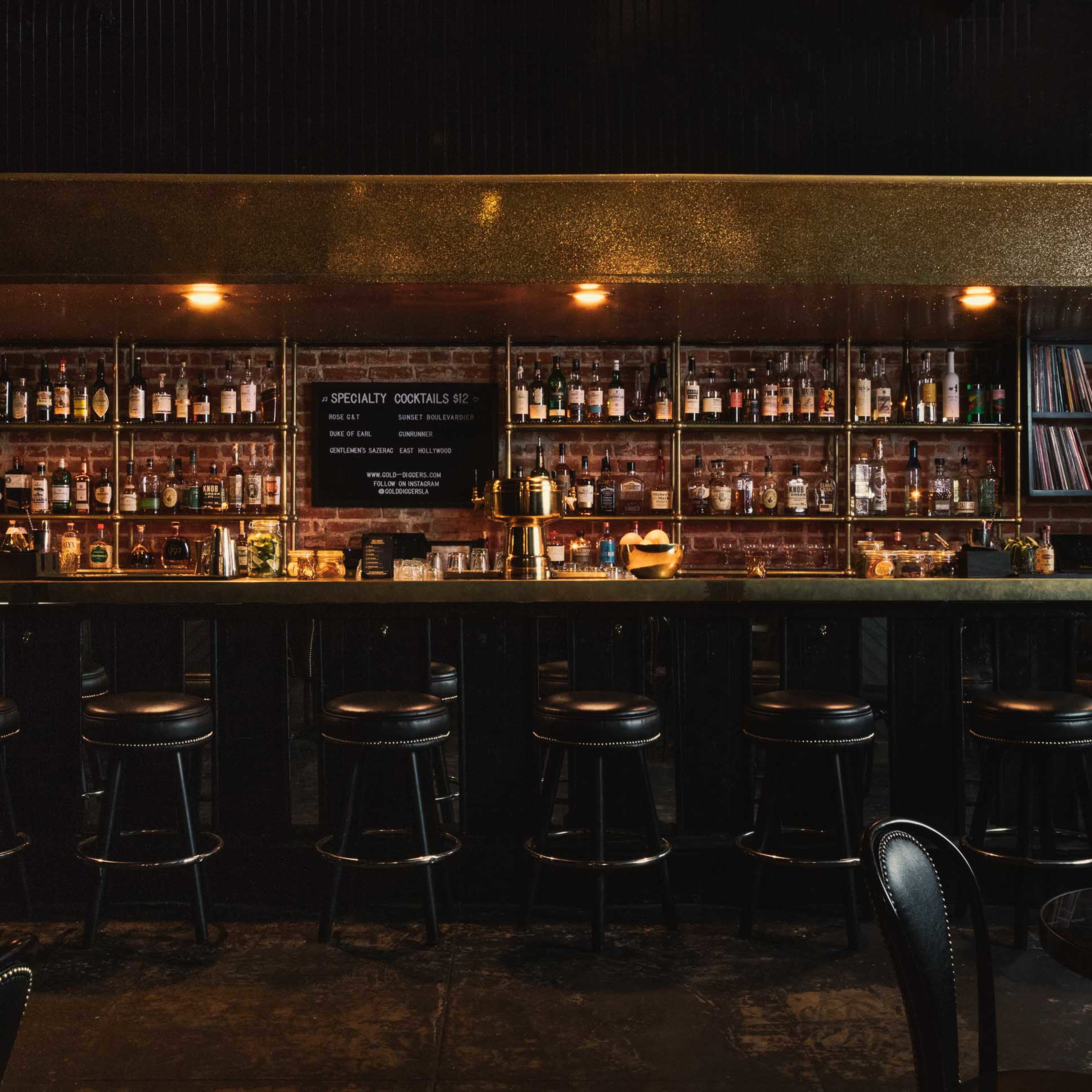 A dimly lit bar with a long counter, featuring shelves filled with various bottles and a few small tables with chairs.