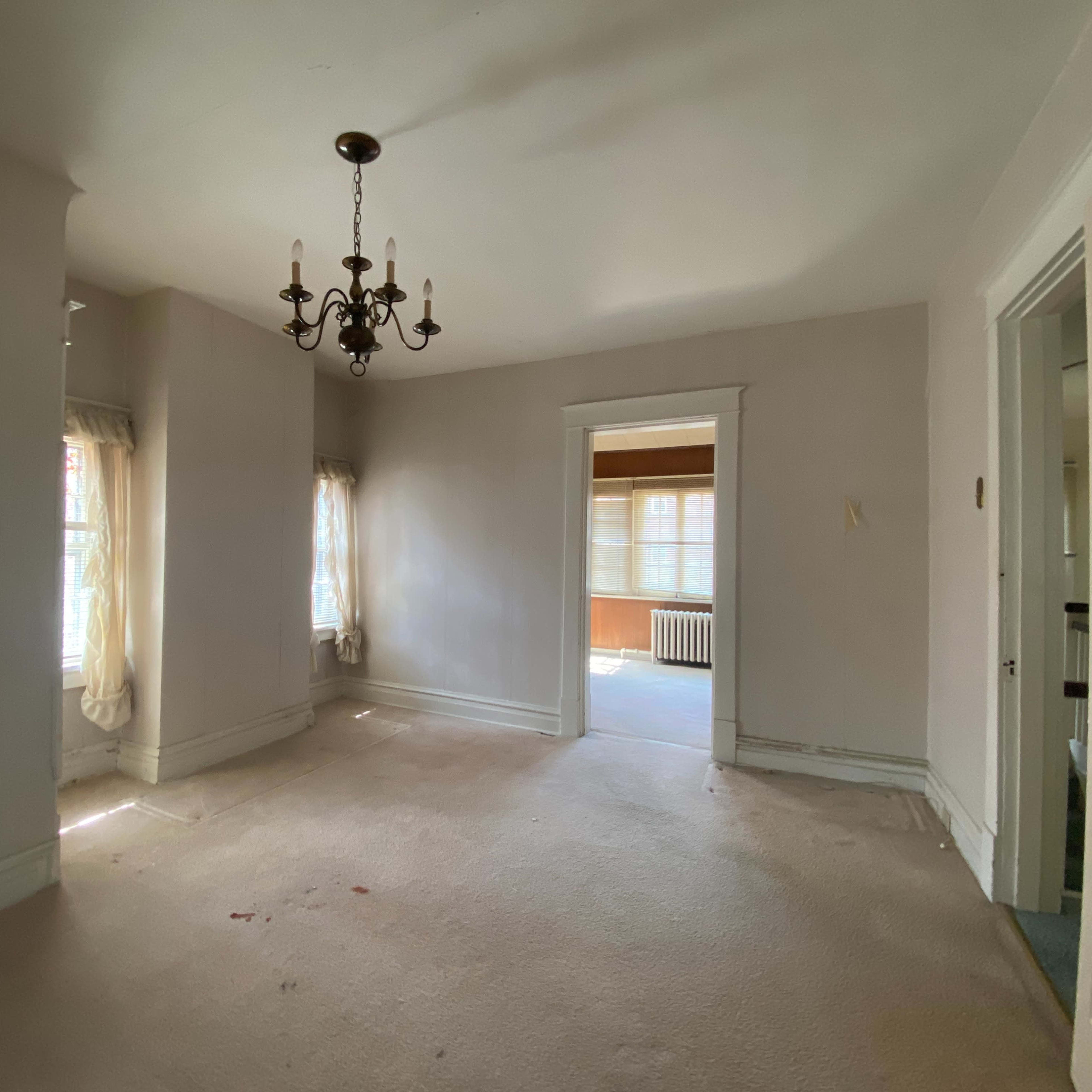 A vacant room features a chandelier, two windows with curtains, and an adjacent room visible through an open doorway.