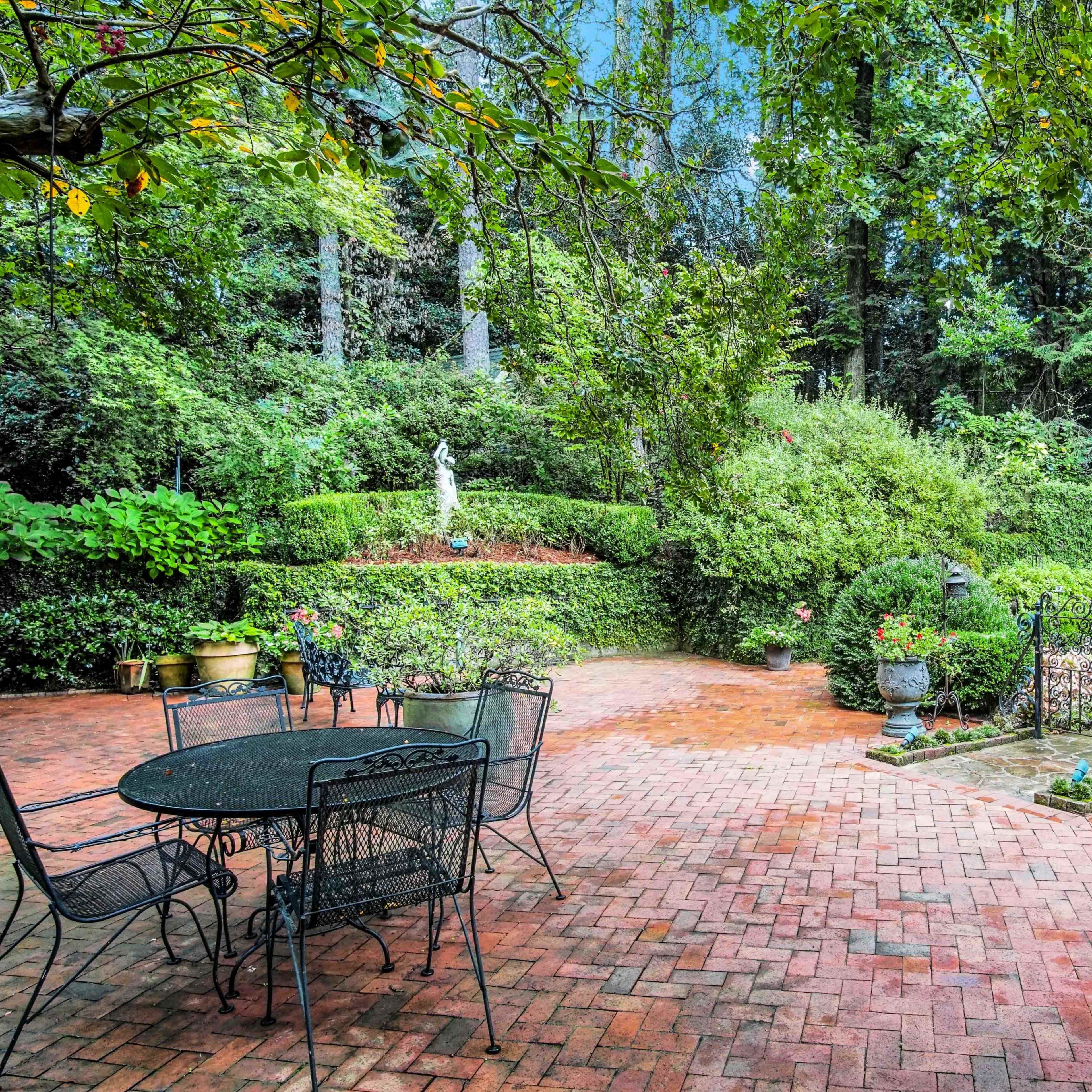 A brick patio surrounded by lush greenery and garden features, including a statue and potted plants.