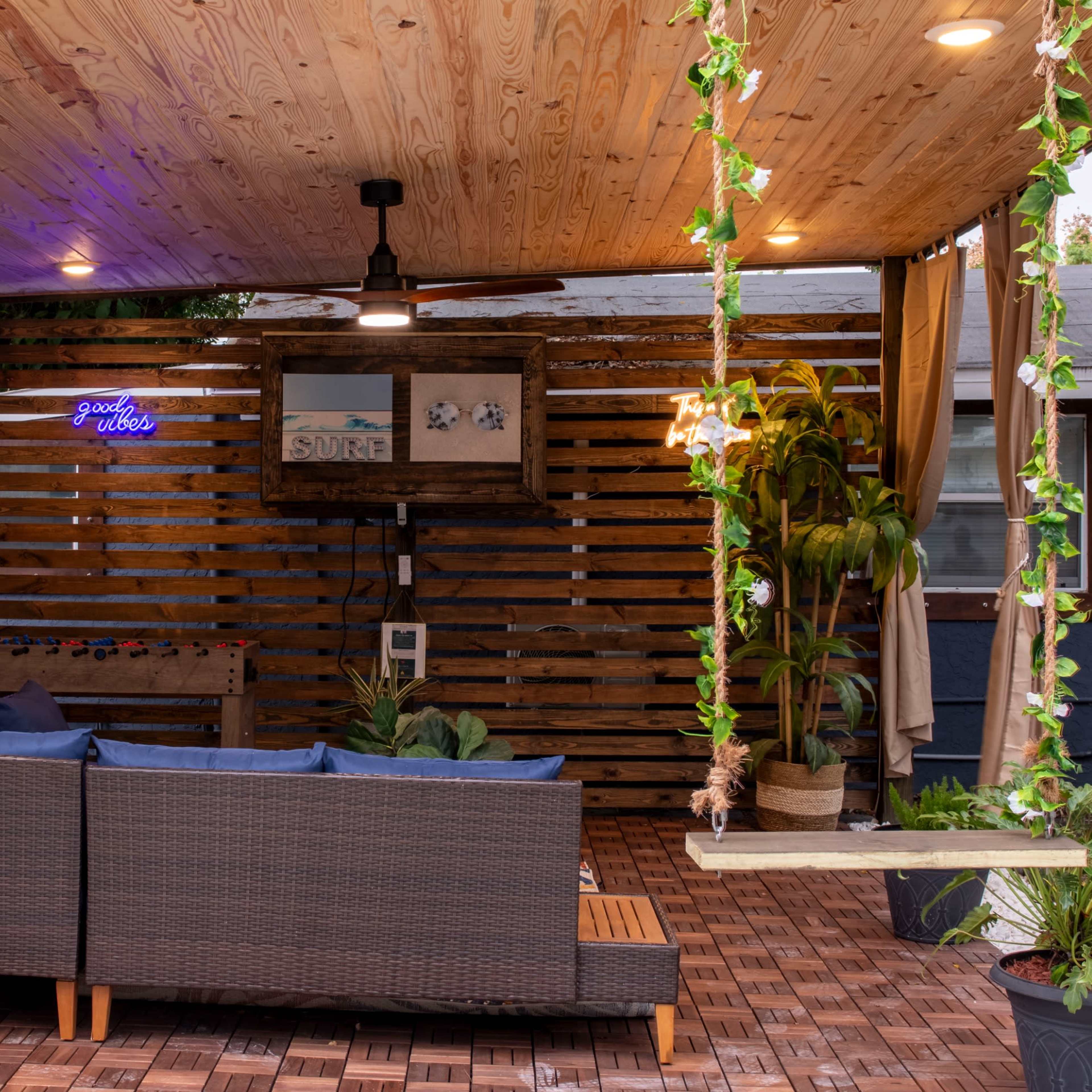 An outdoor seating area with a wooden ceiling, a wall mural of a beach scene, and a brown couch adorned with blue cushions, surrounded by plants and string lights.