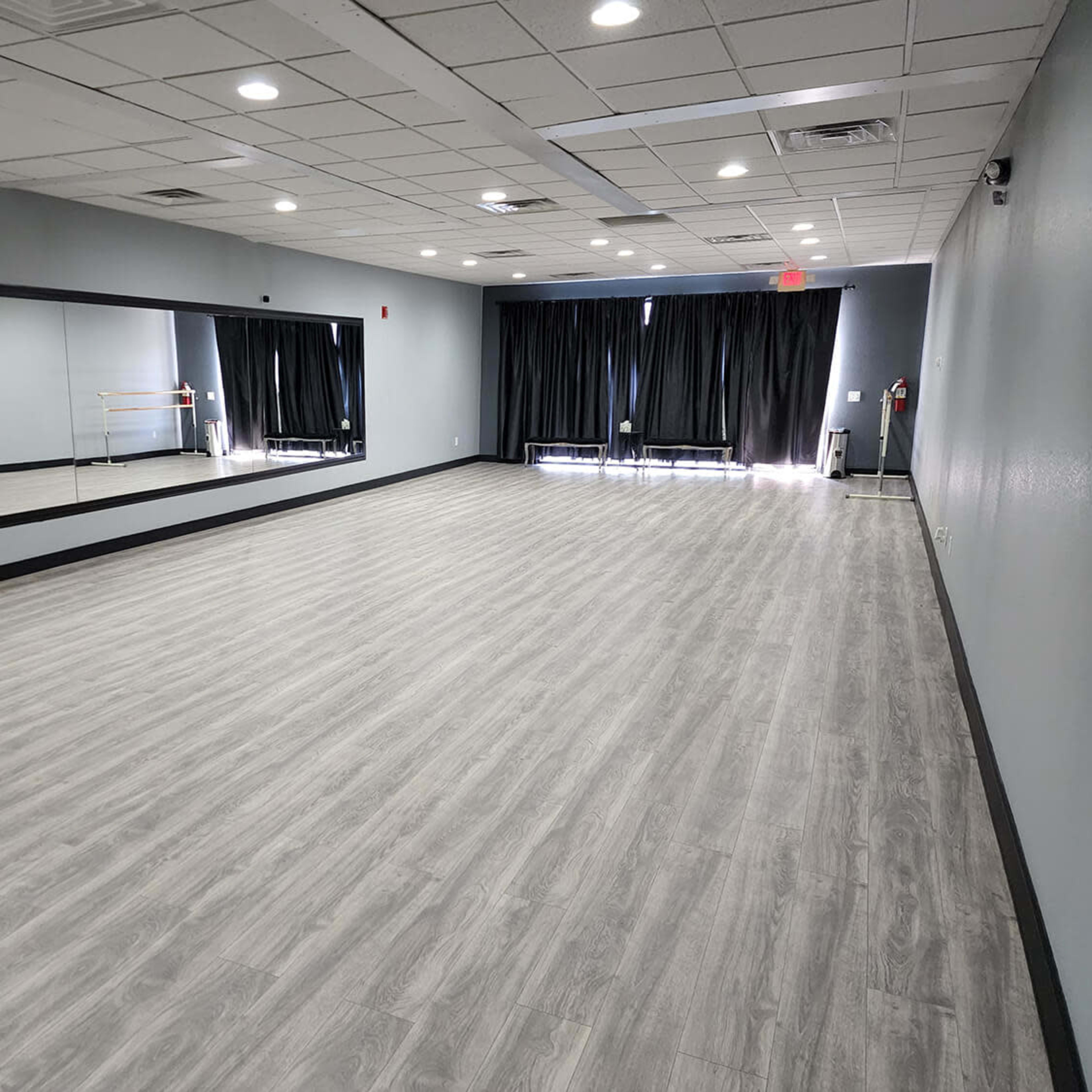 A spacious dance studio with gray walls, a large mirror on one side, and black curtains covering the windows.
