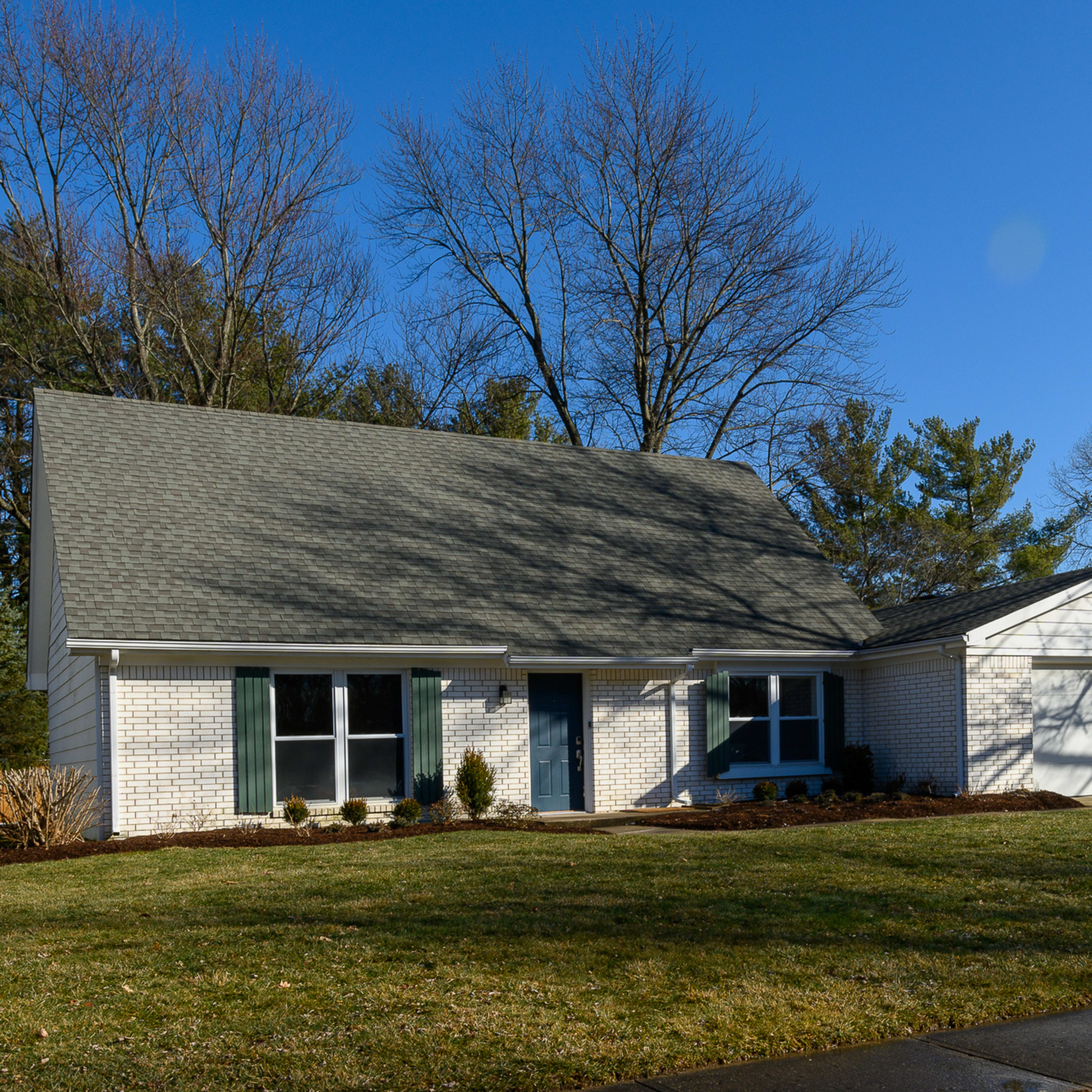 A single-story house with a gabled roof, green shutters, and a two-car garage is situated on a lawn surrounded by trees.