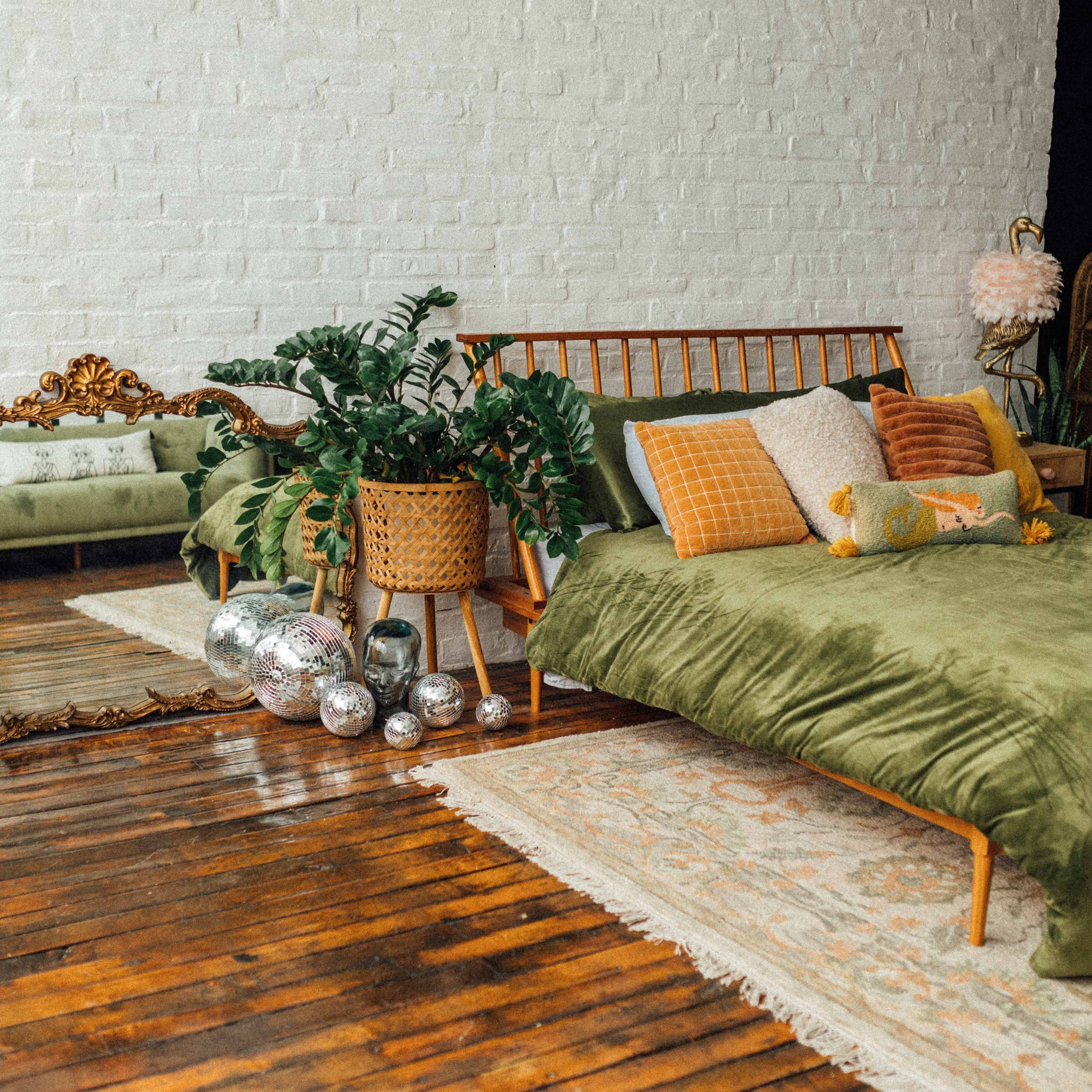 The scene features a cozy bedroom with a green bedding set, a wooden bed frame, a vintage-style mirror, and decorative plants, all arranged on a wooden floor with a patterned rug.