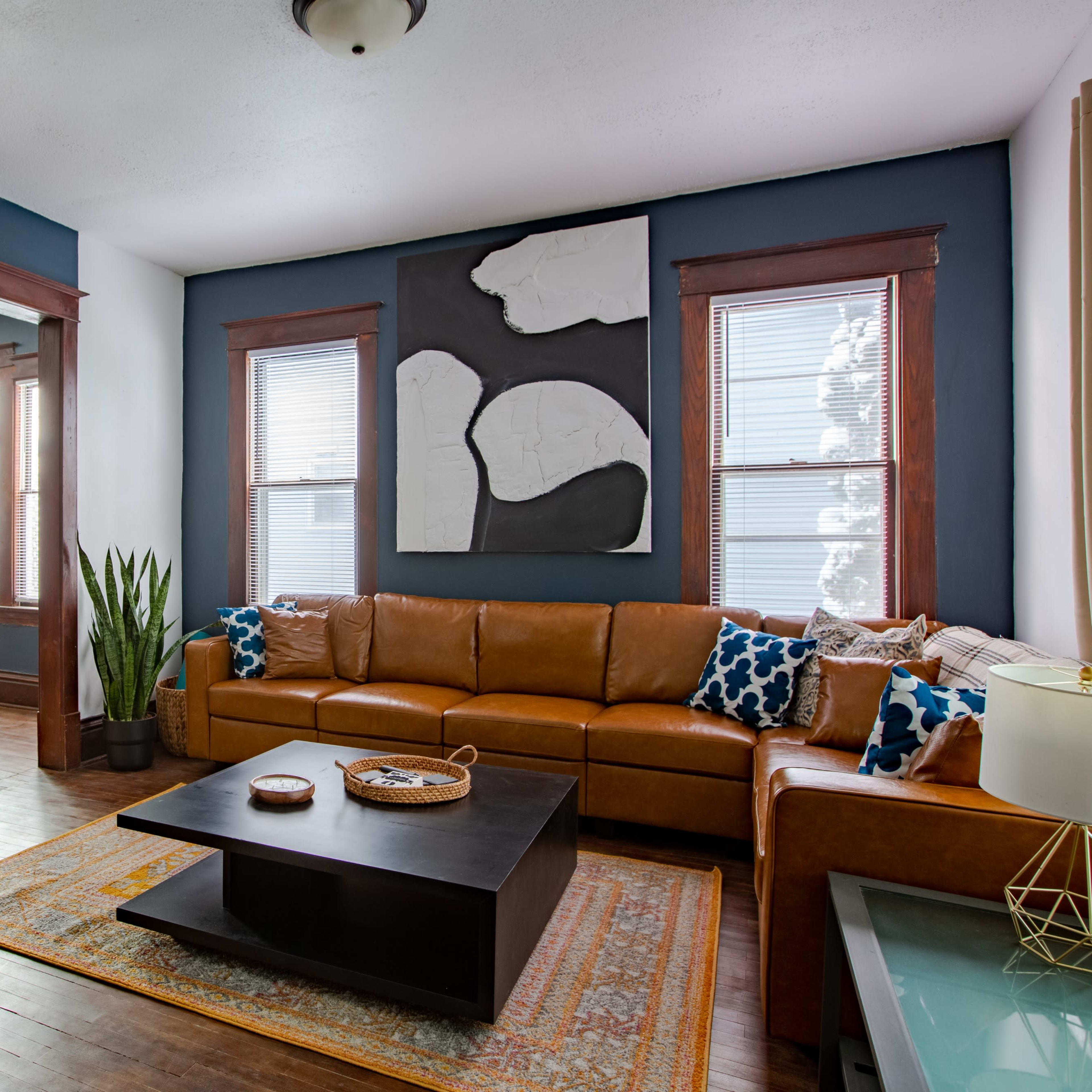A living room with a brown leather sectional sofa, a black coffee table, and a large abstract wall decoration, with a dining area visible through an archway.