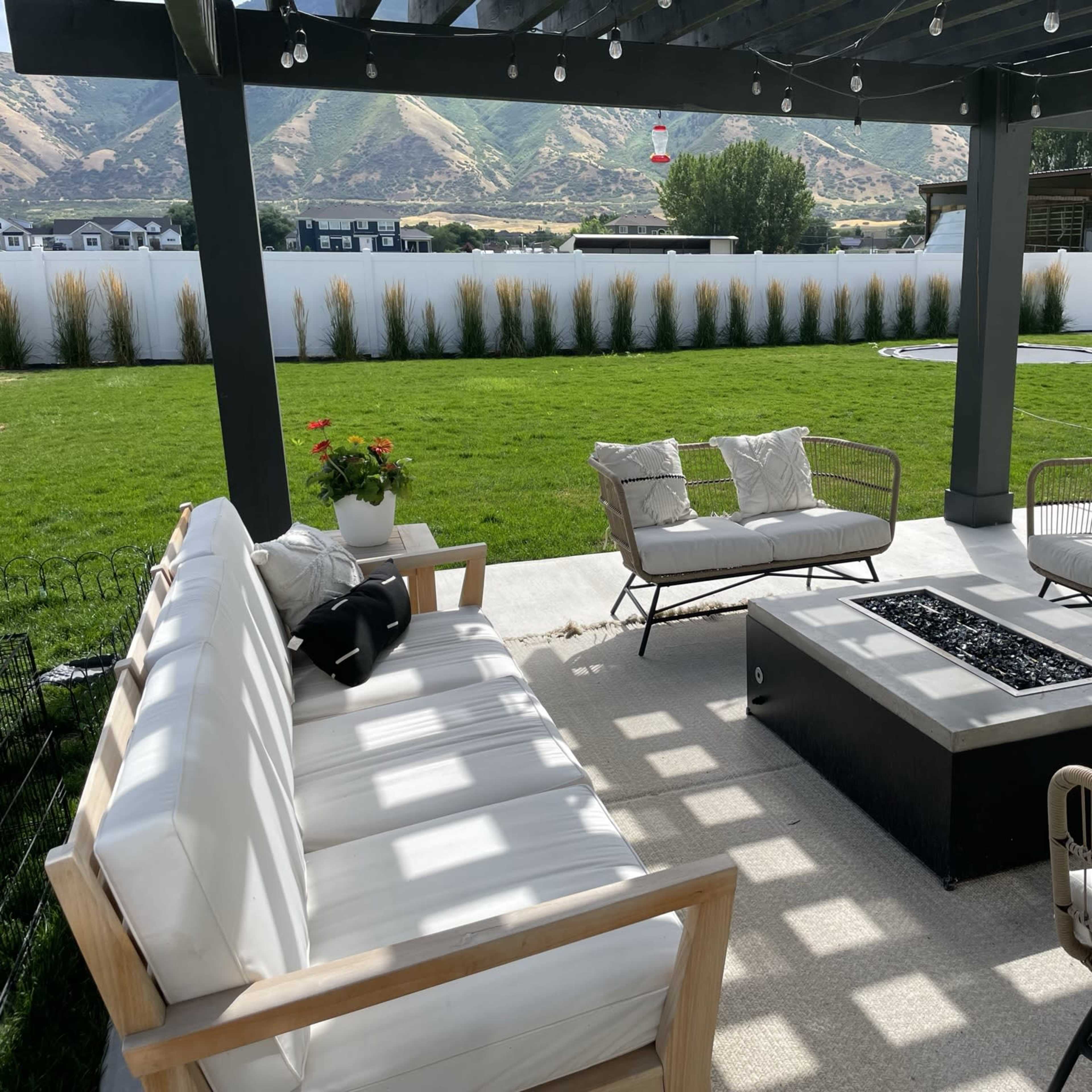 A covered patio features a seating arrangement around a fire pit, set against a backdrop of a green lawn and mountains.