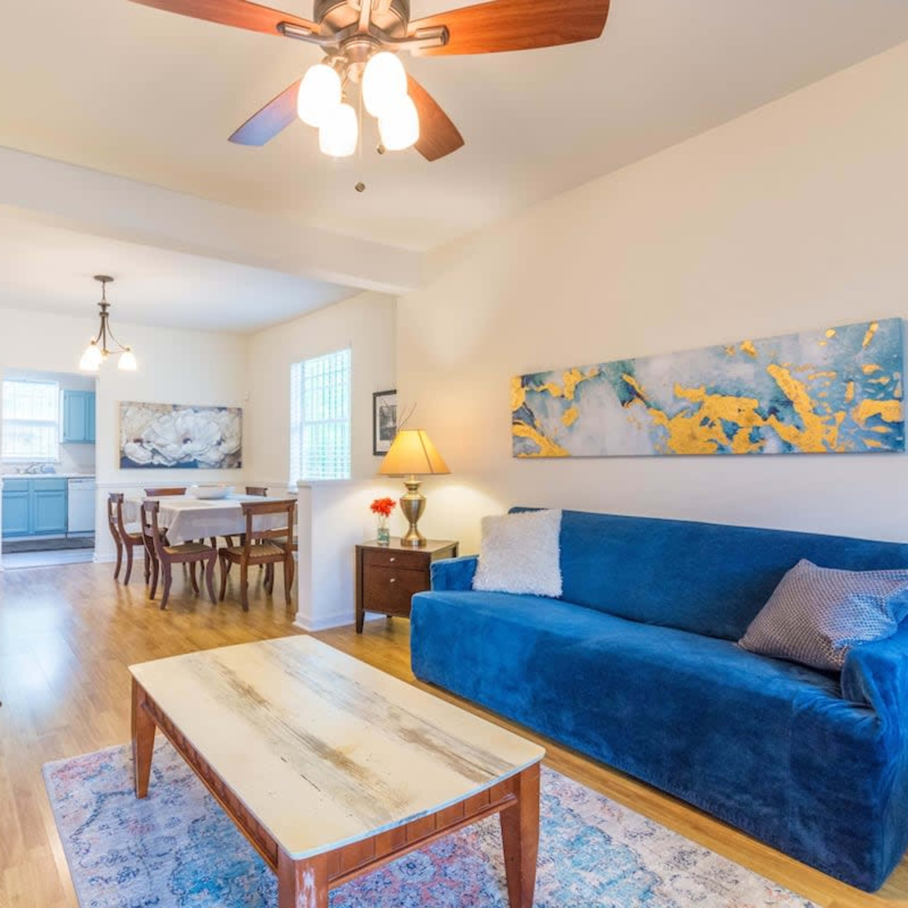 A bright living room with a blue sofa, a wooden coffee table, and a dining area in the background.