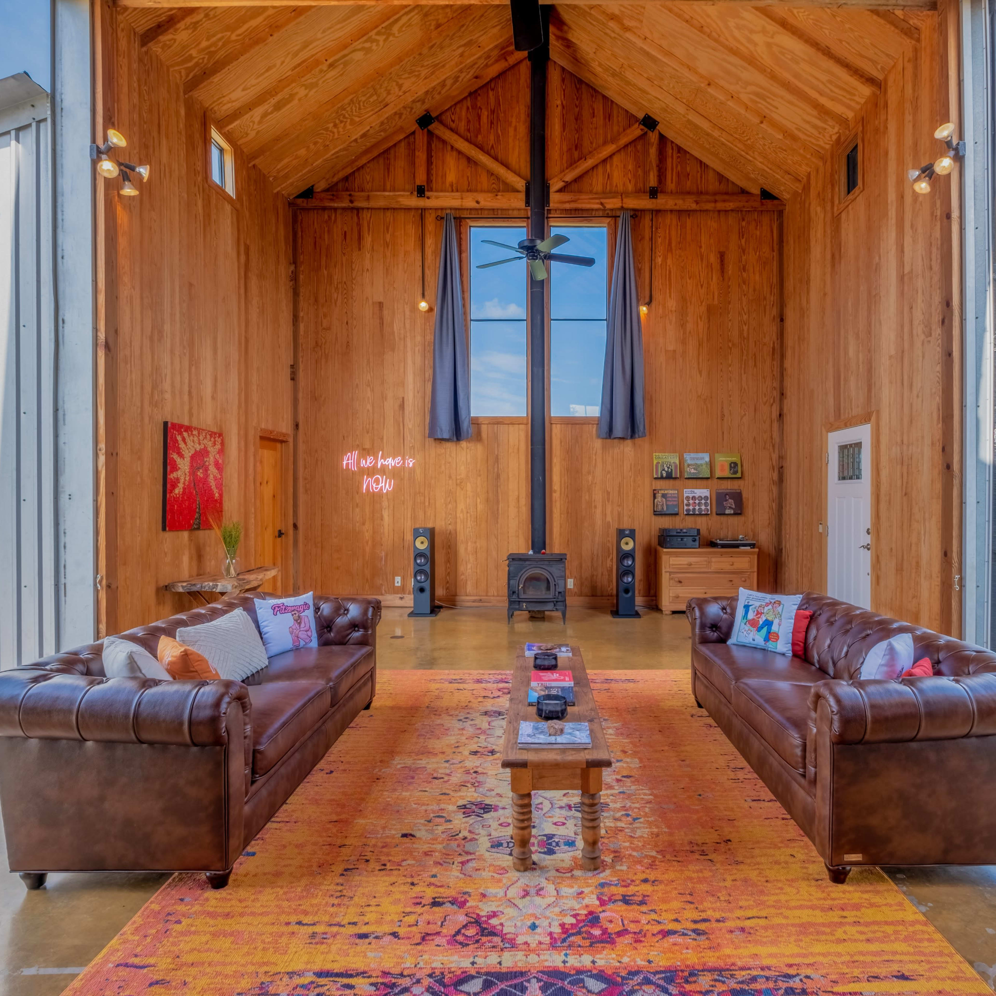 A spacious, rustic interior features two leather sofas facing a central coffee table on a vibrant rug, with large windows allowing natural light to fill the room.