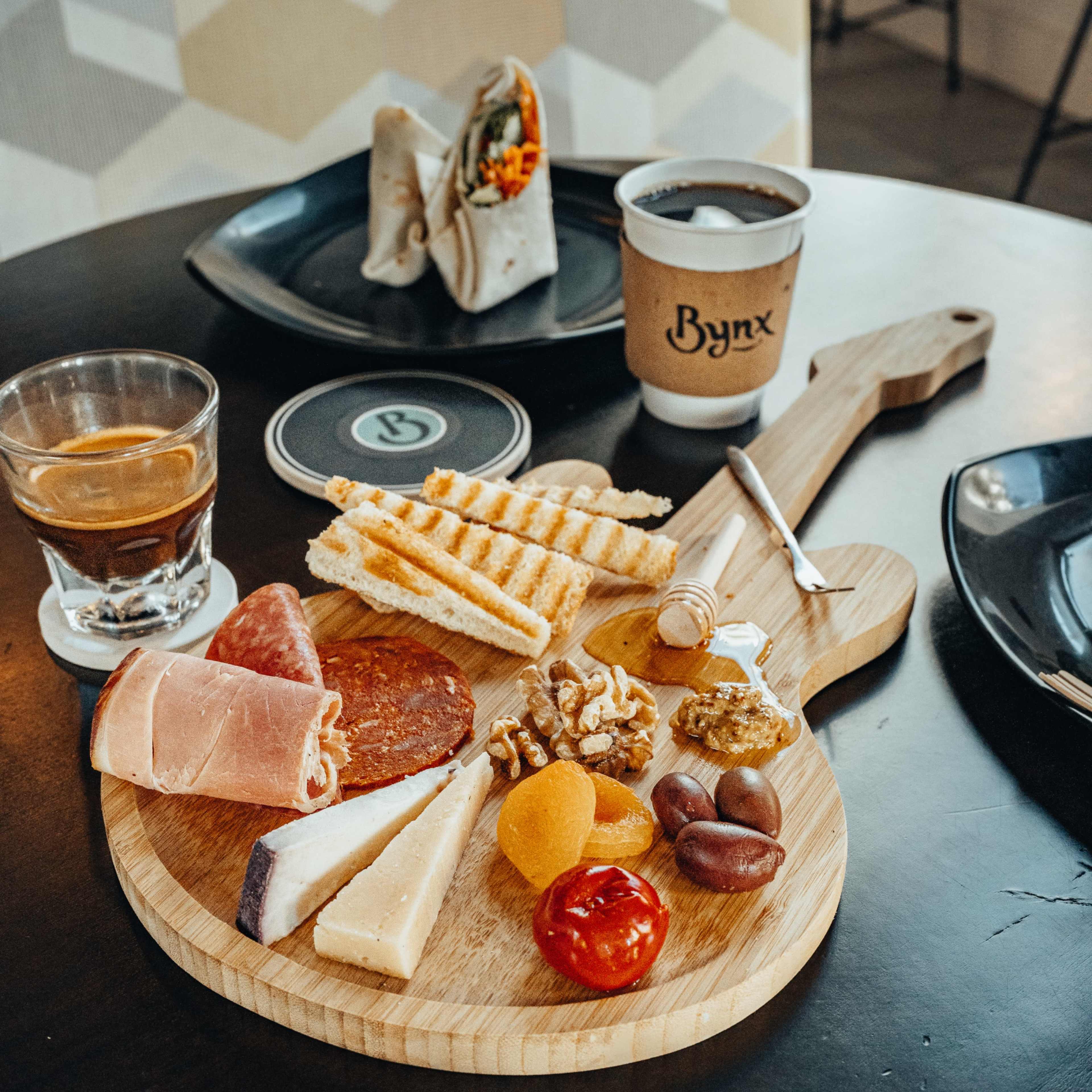 A wooden platter with assorted cheeses, meats, nuts, dried fruits, and toasted sandwiches, accompanied by a glass of whiskey and a coffee cup on a dark table.