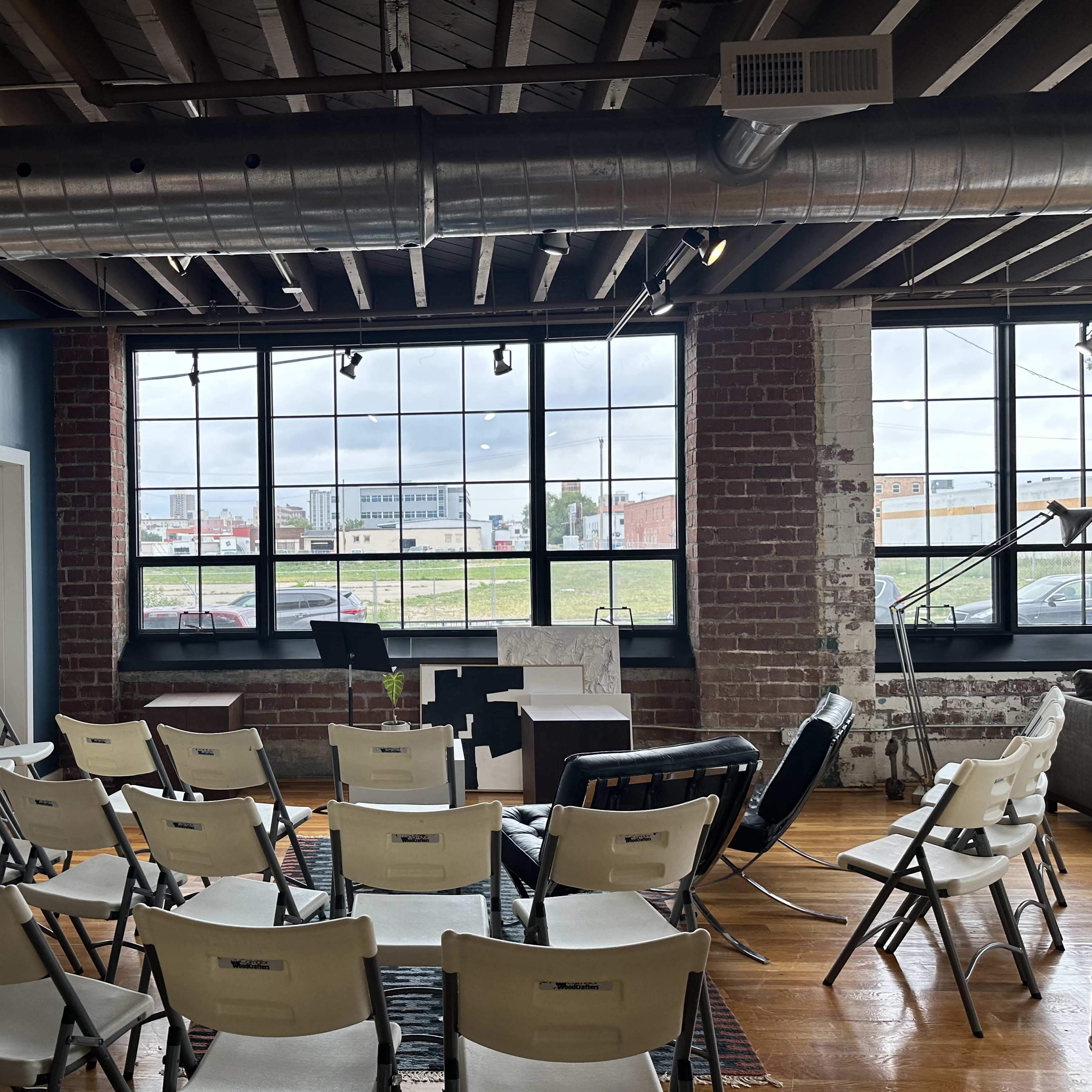 A room with rows of white folding chairs facing a small stage, surrounded by large windows that view an outdoor area with grassy fields and buildings.