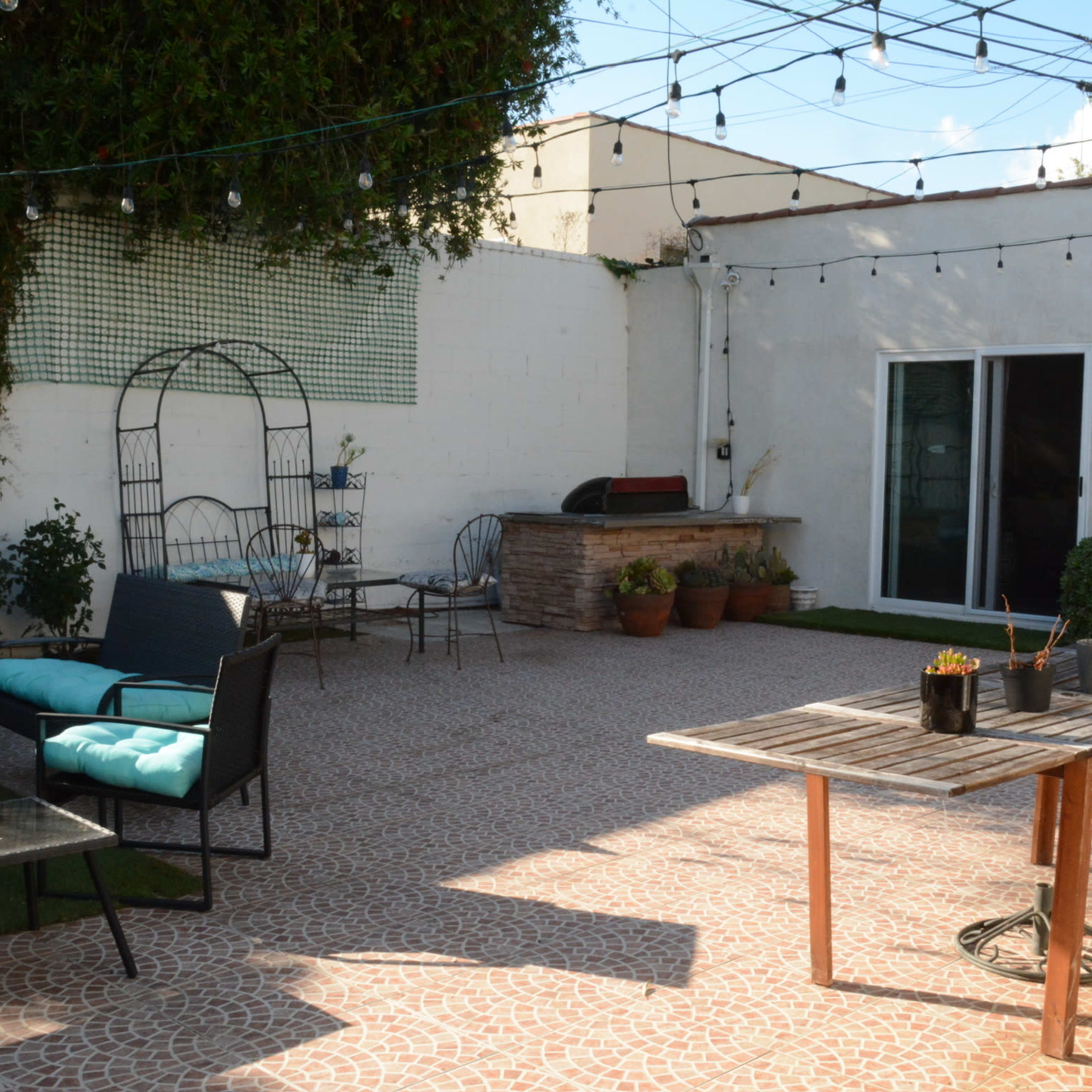 The outdoor space features a paved patio with seating arrangements, a wooden table with potted plants, and string lights overhead.