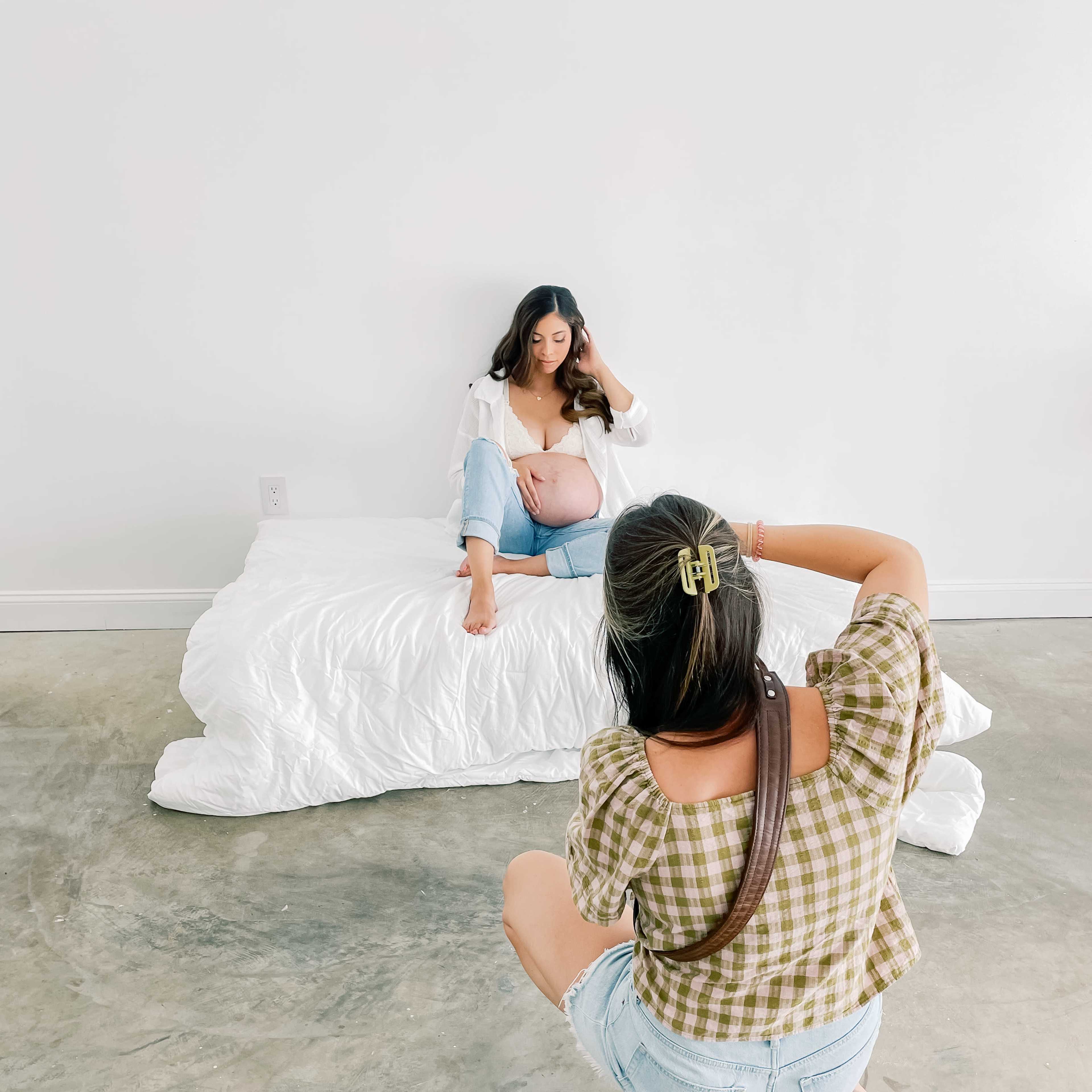 A woman sitting on a white bed is posing for a photograph while another woman crouches behind the camera.