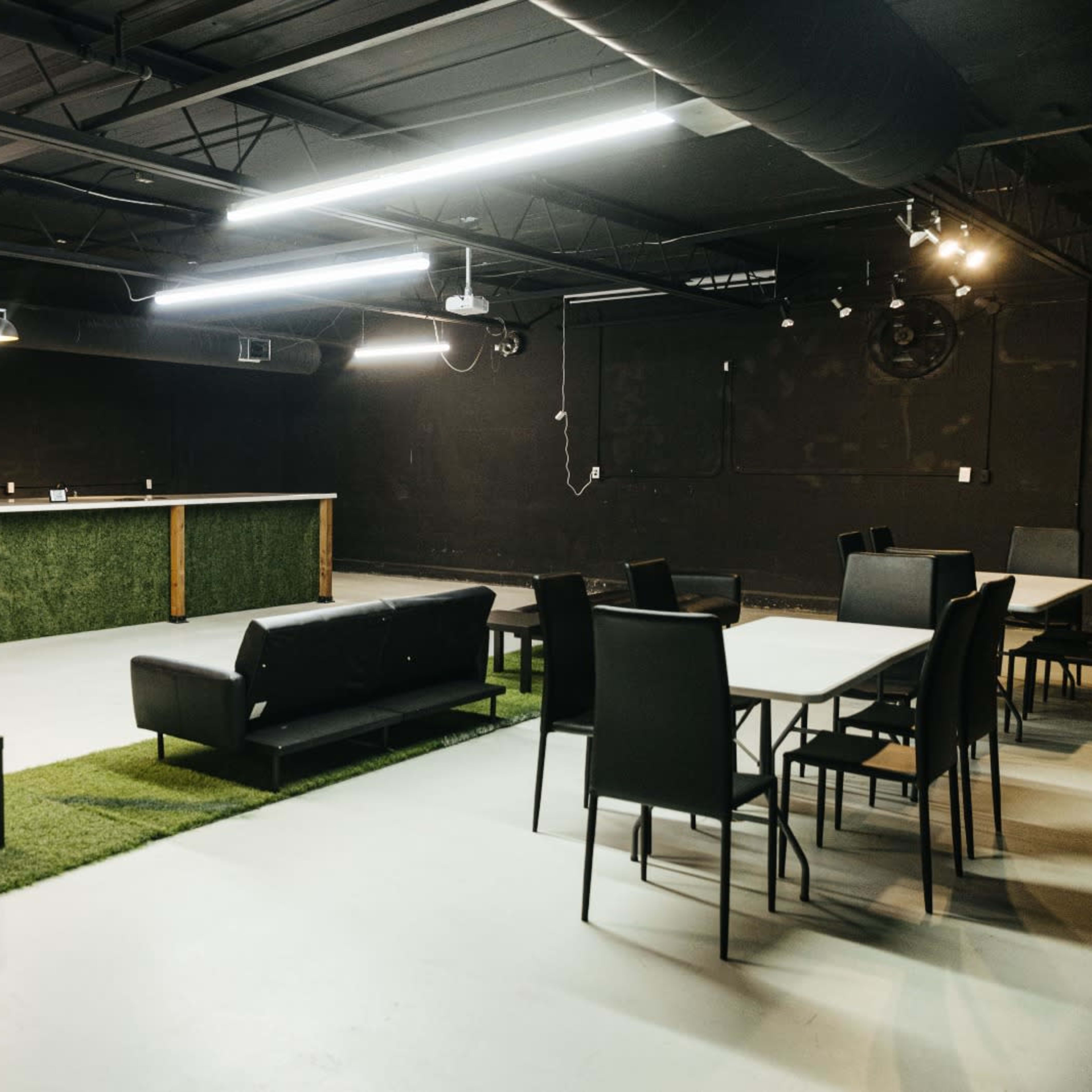 A spacious, modern lounge area featuring dark walls, a bar with a green backdrop, several black seating options, and tables set on a light-colored floor.