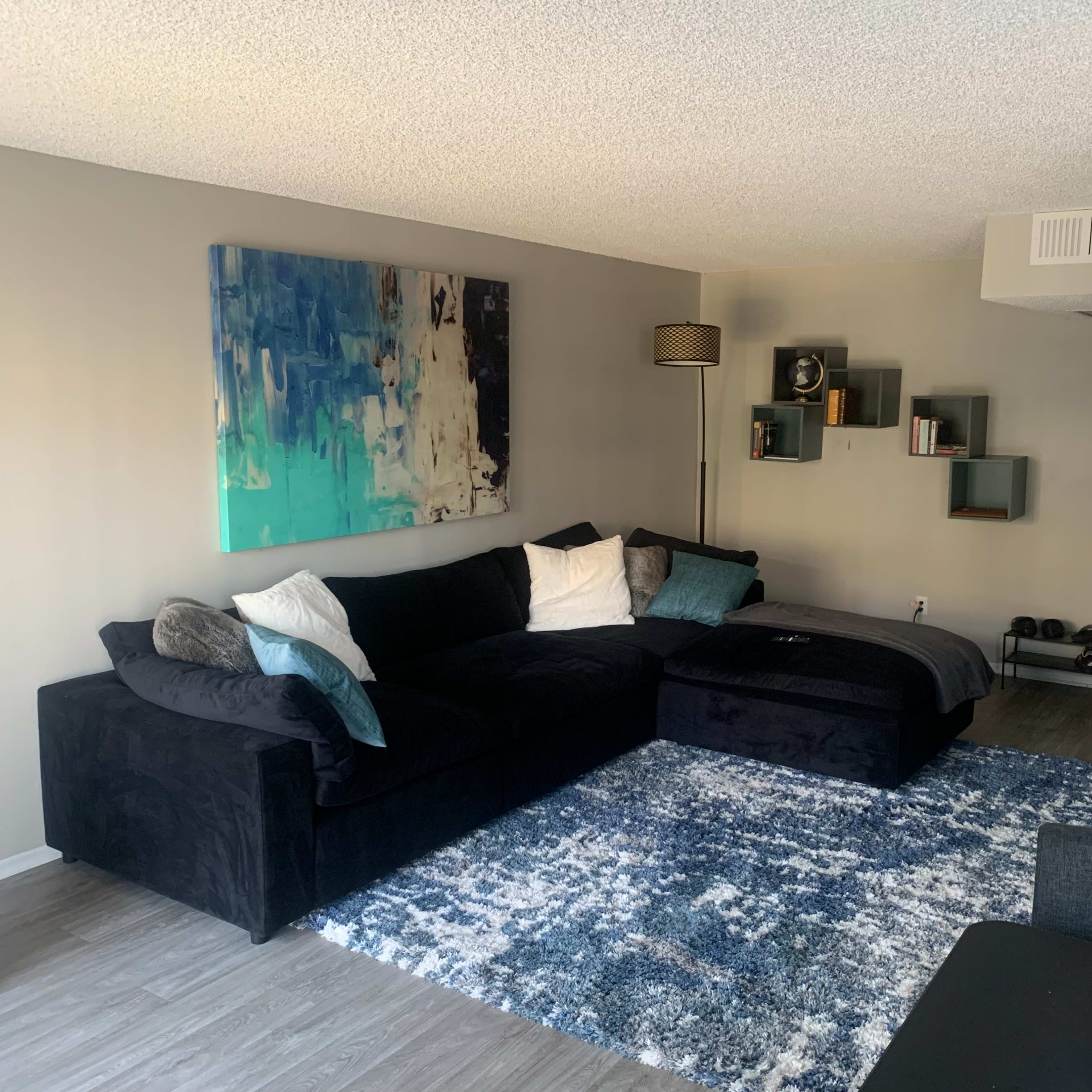 A modern living room with a black sectional sofa, a blue abstract painting on the wall, and a bookshelf with decorative items.