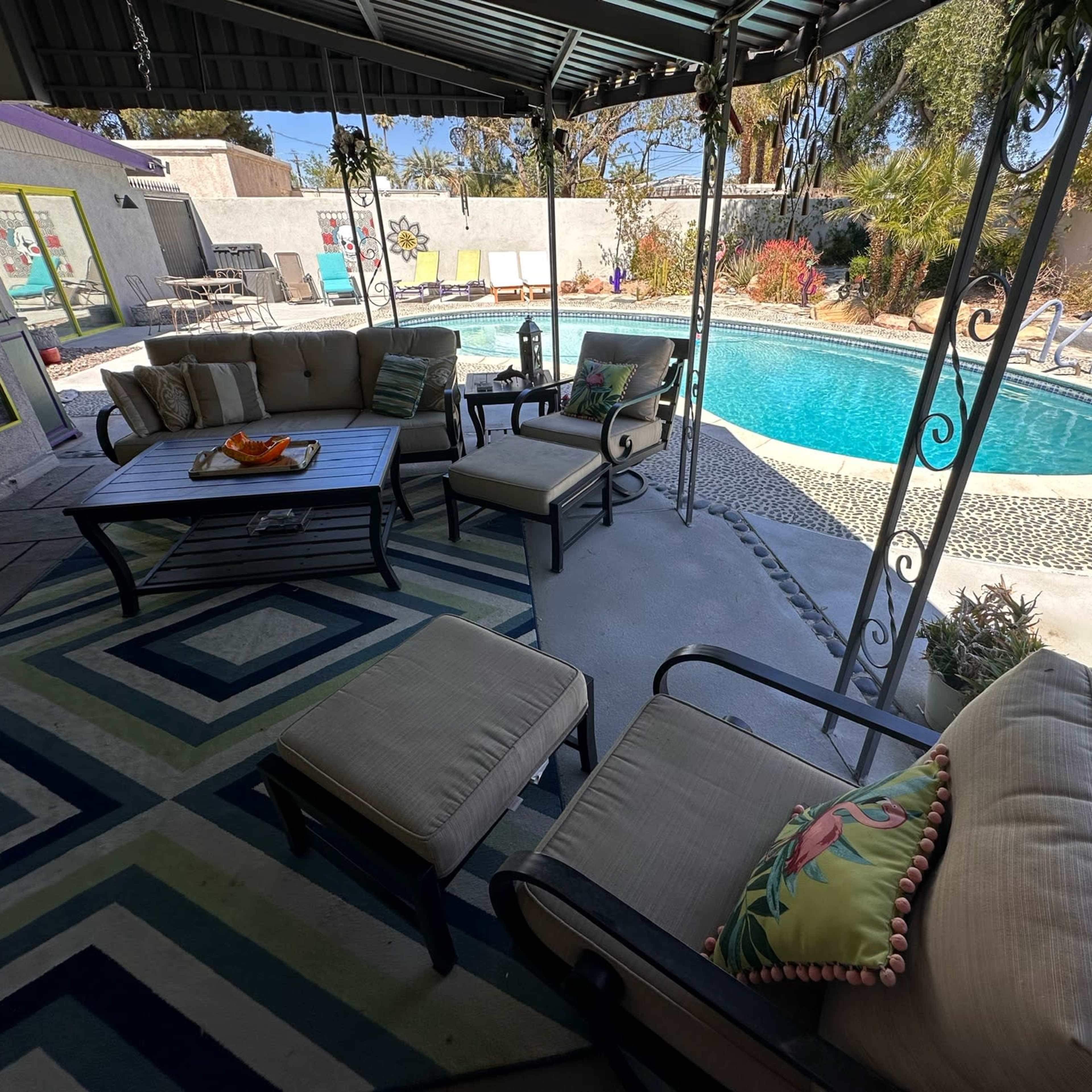 A covered patio with seating arranged around a table, overlooking a swimming pool surrounded by landscaped areas.