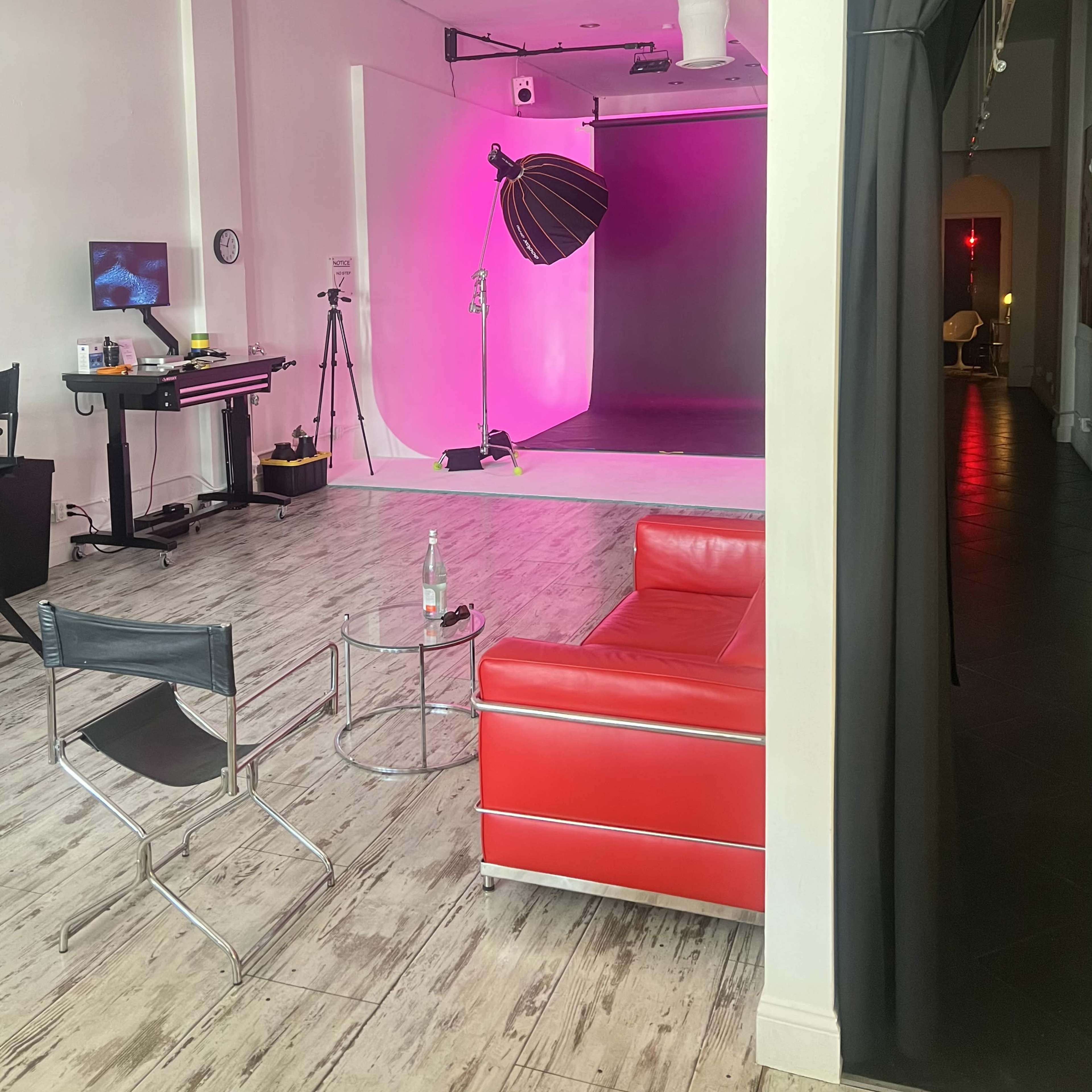 A studio space featuring a red couch, a lighting setup with a softbox, and a pink backdrop along with various equipment.