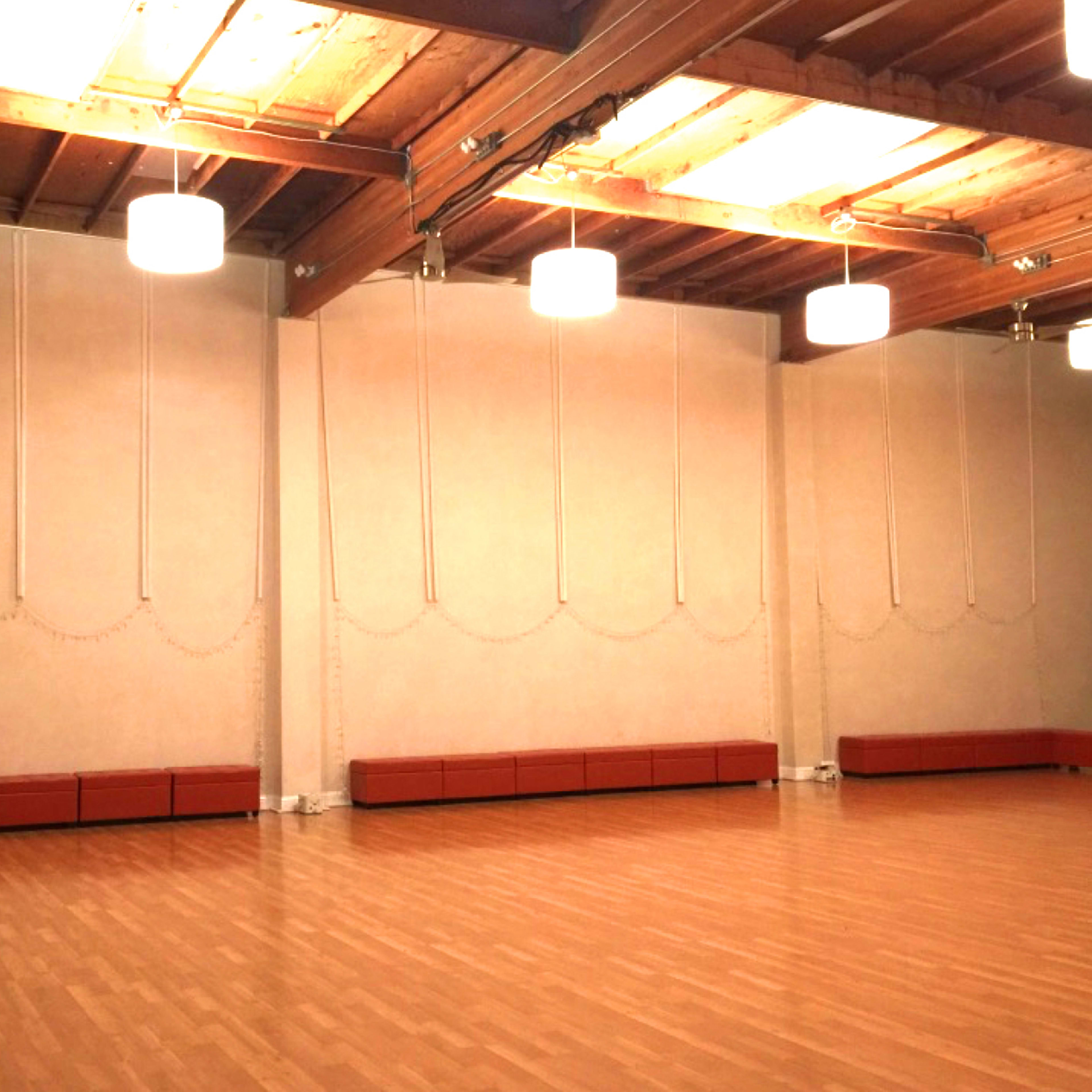 A spacious indoor room with wooden floors, wall-mounted lights, and a long padded bench along one wall.