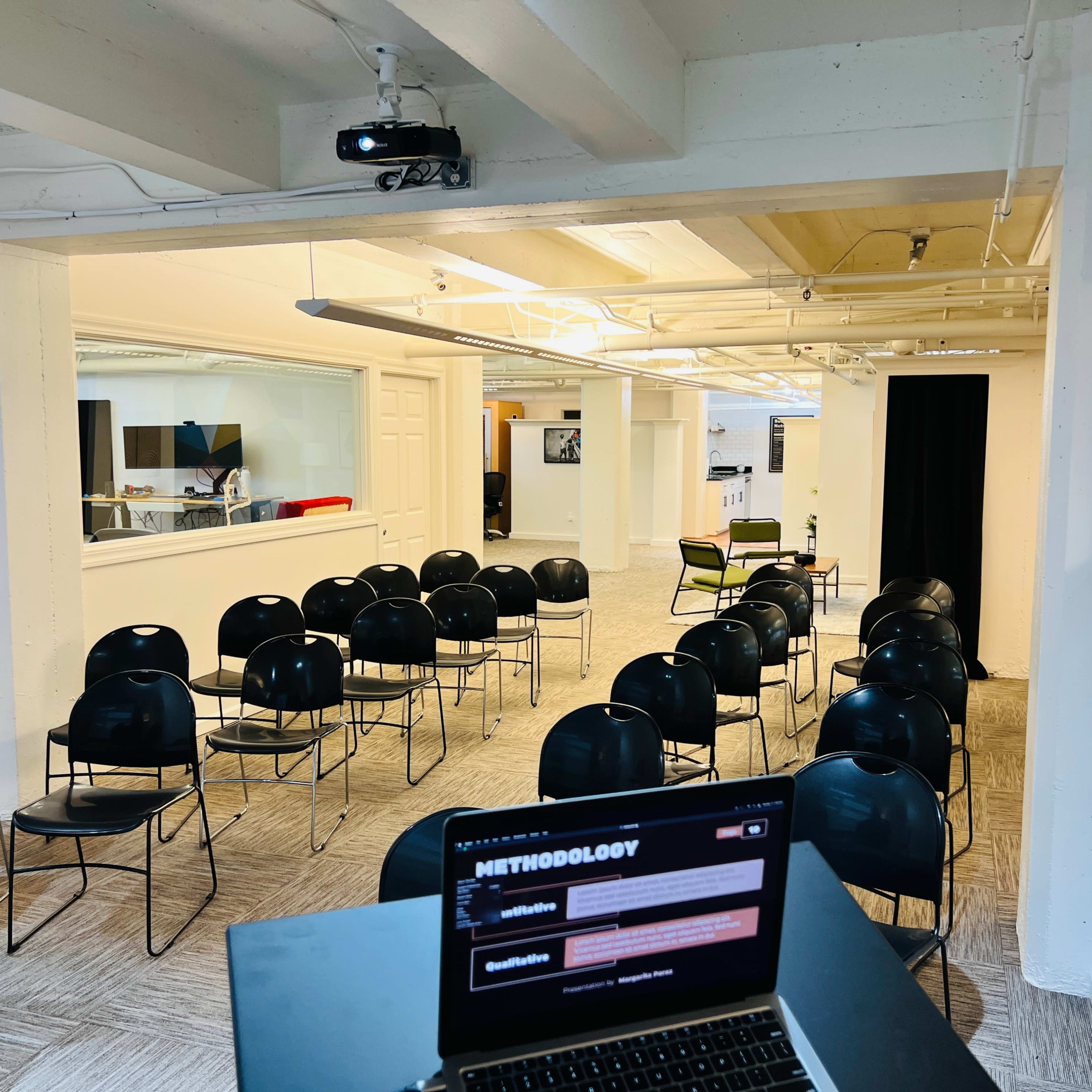 A spacious room with rows of black chairs facing a projector screen and a laptop on a table in the foreground.