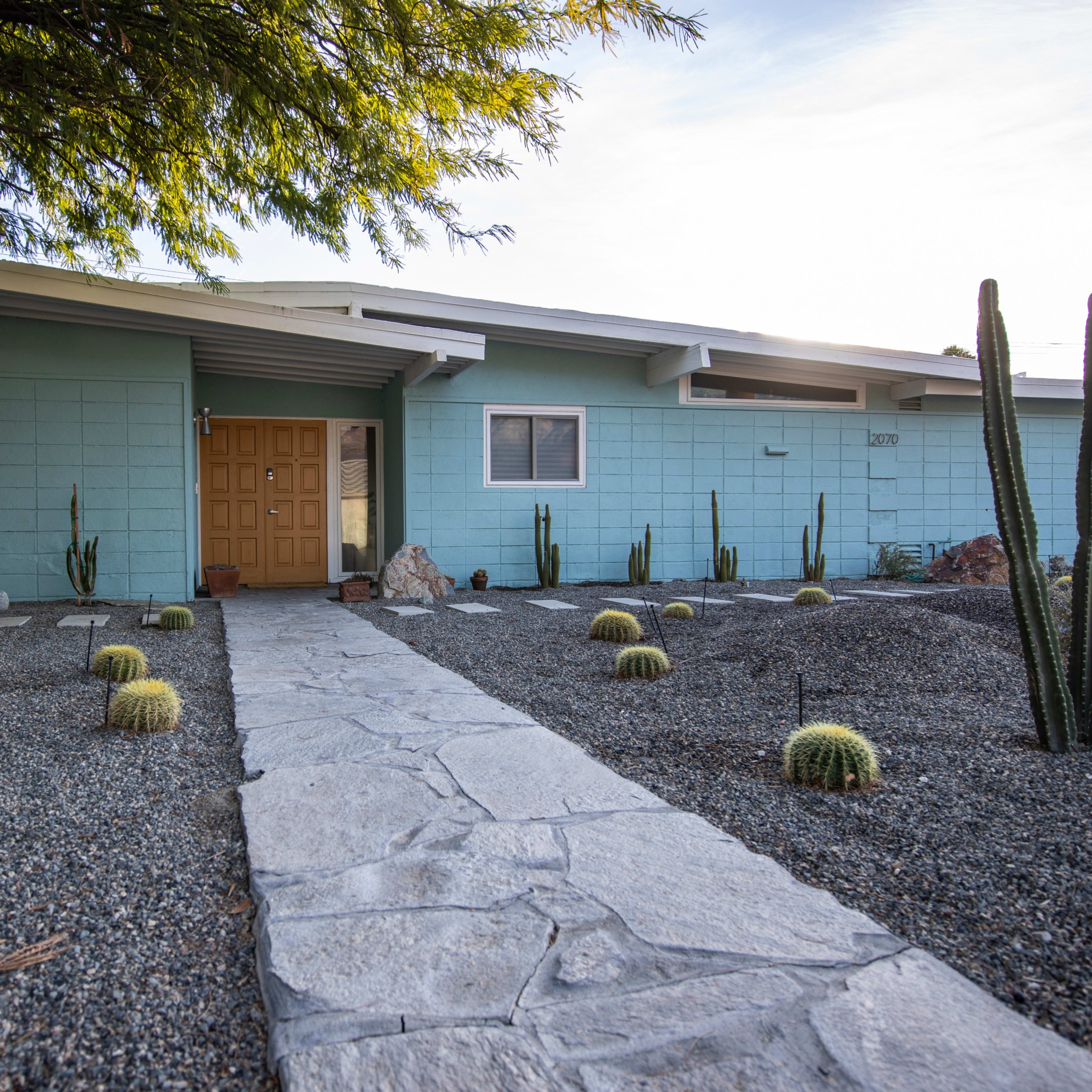 A modern home with a light blue exterior and a stone pathway leading to a wooden door, surrounded by a landscaped area featuring cacti and gravel.