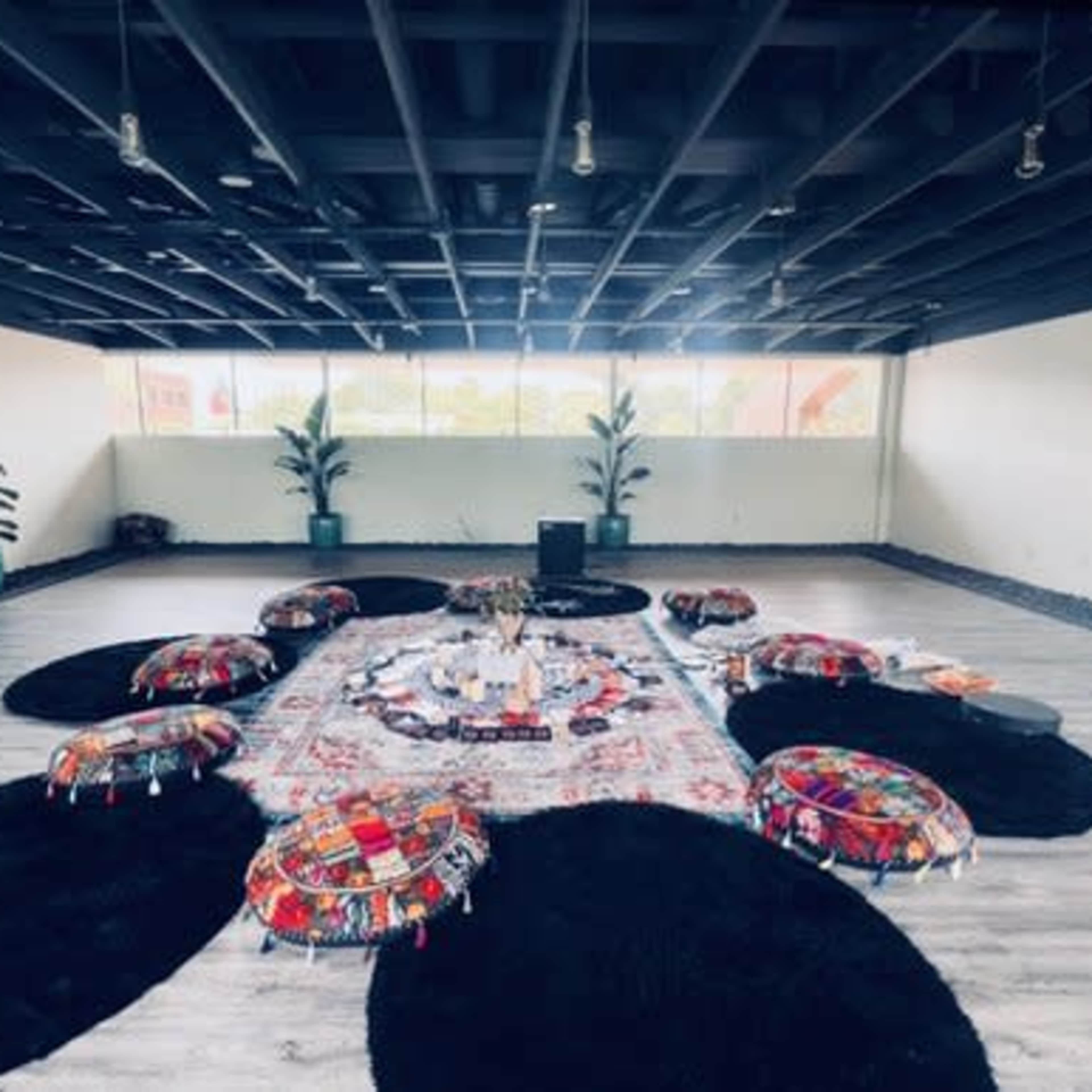 A spacious, minimalist room with a centrally arranged area featuring colorful poufs and a decorative rug surrounded by plants.