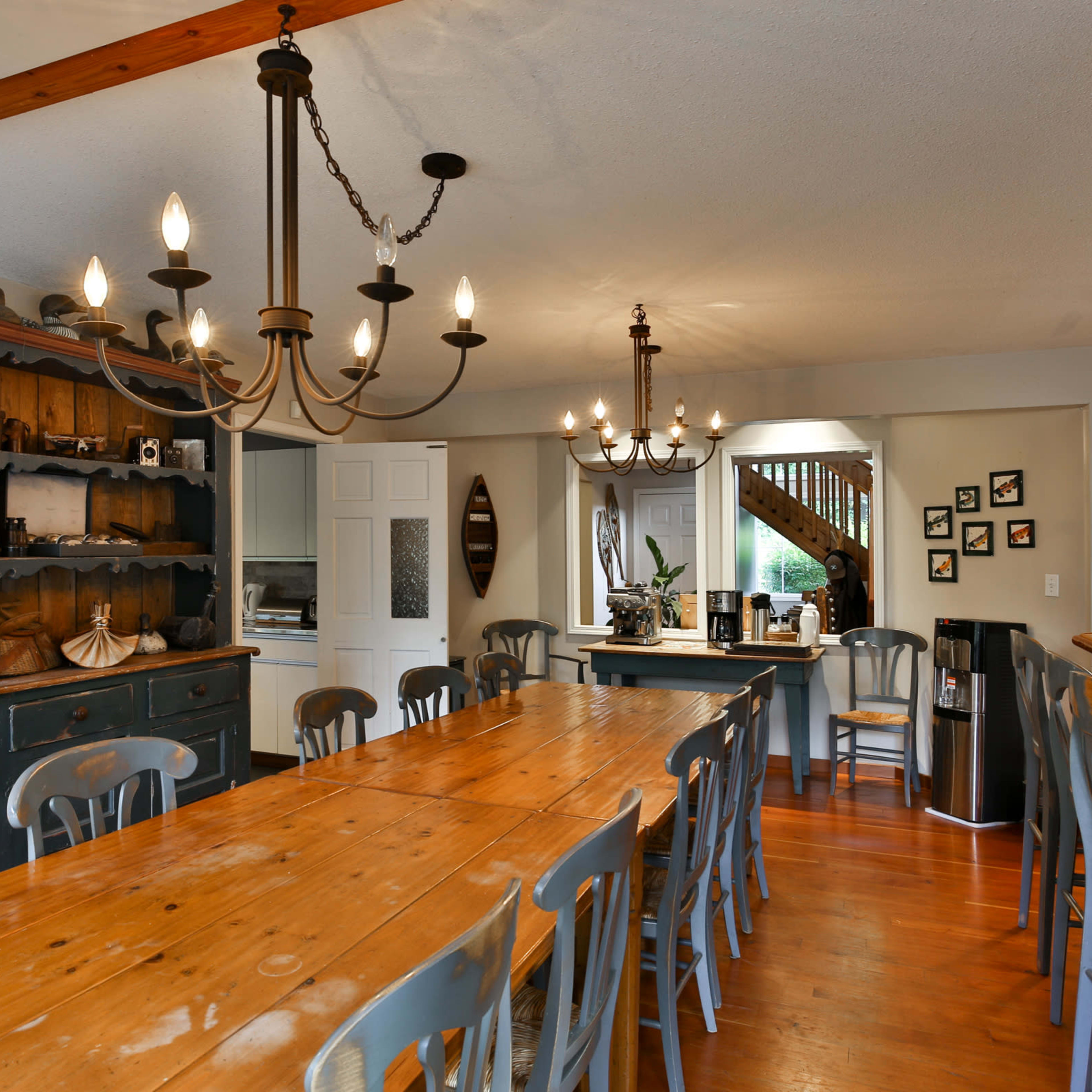 A spacious dining area with a long wooden table surrounded by chairs, accented by two stylish chandeliers and a sideboard filled with decorative items.