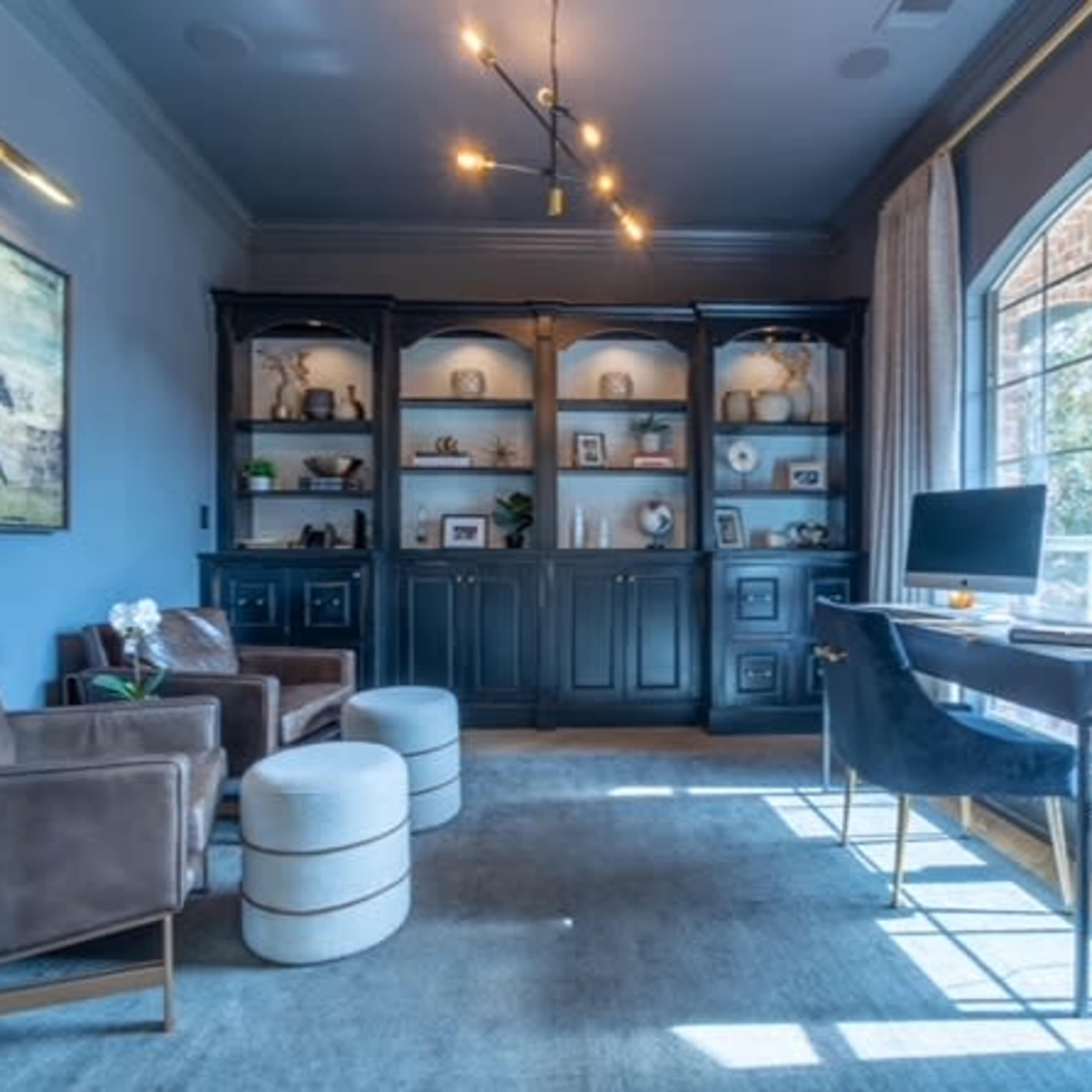A well-lit home office features a desk, shelving unit, and seating area with two armchairs and an ottoman.
