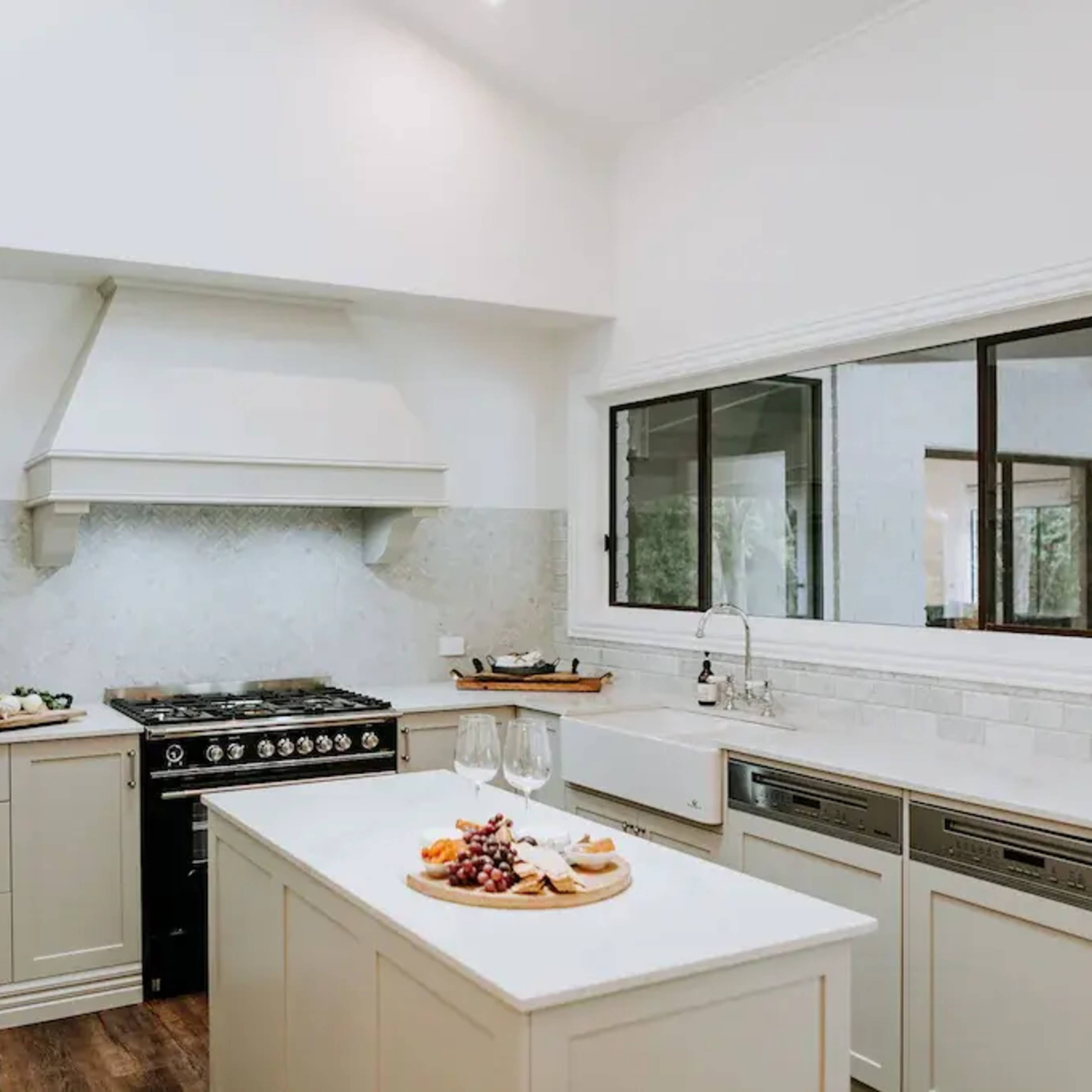A modern kitchen featuring a central island with wine glasses and a cheese platter, a large stove, and a window overlooking the outside.