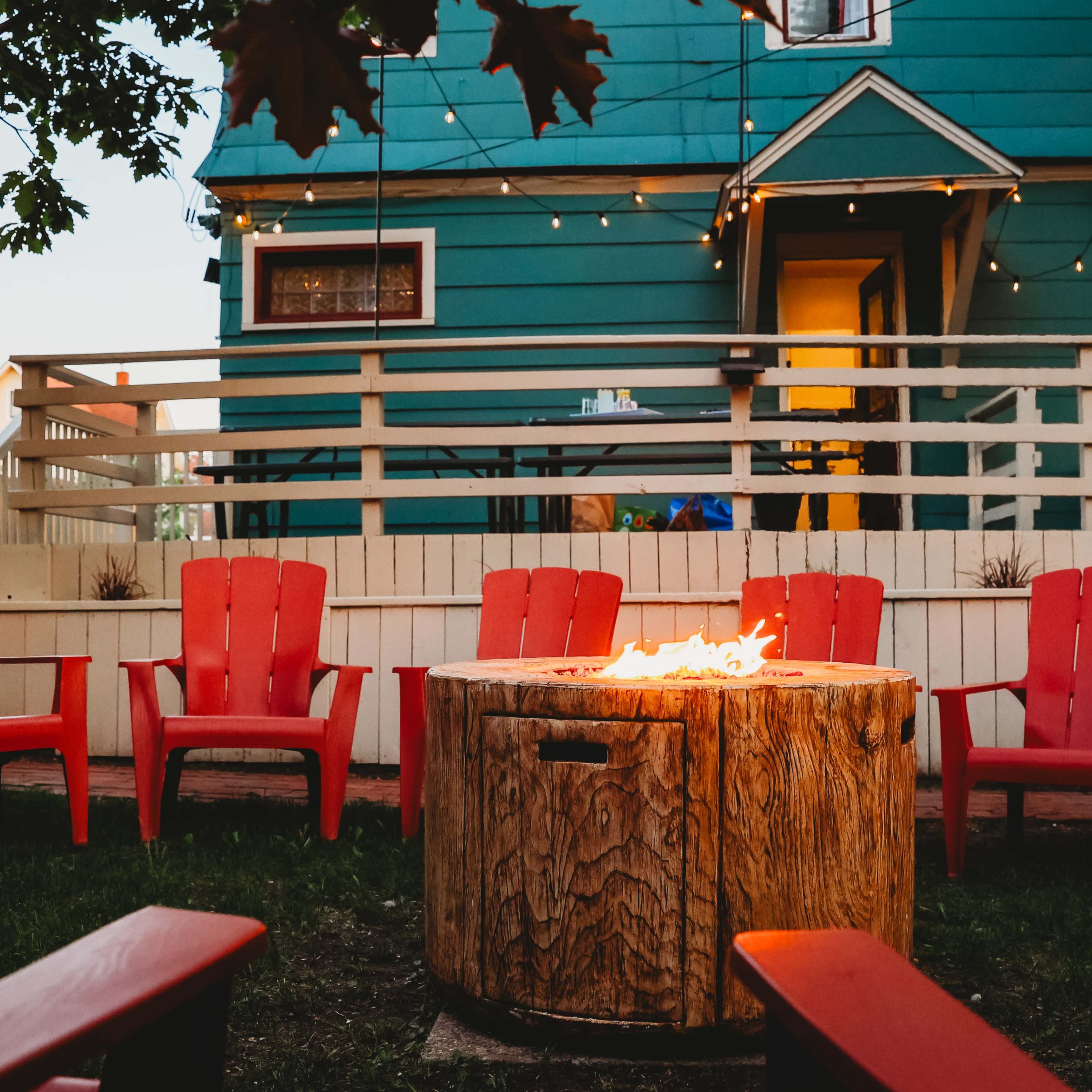 A circular fire pit surrounded by red Adirondack chairs in a backyard with a teal house in the background and string lights overhead.
