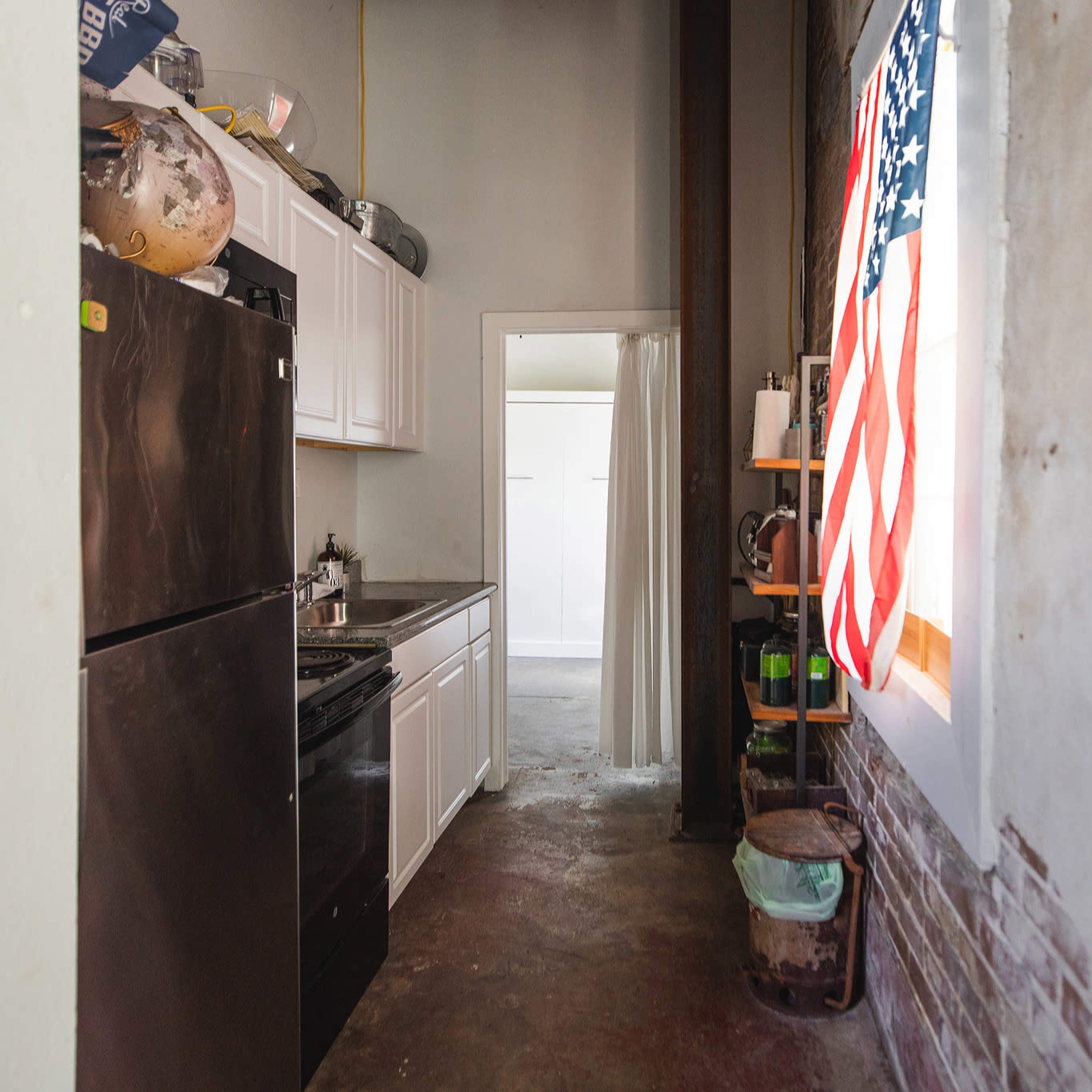 A narrow kitchen area with white cabinetry, a black stove and refrigerator, and an American flag hanging by a window.