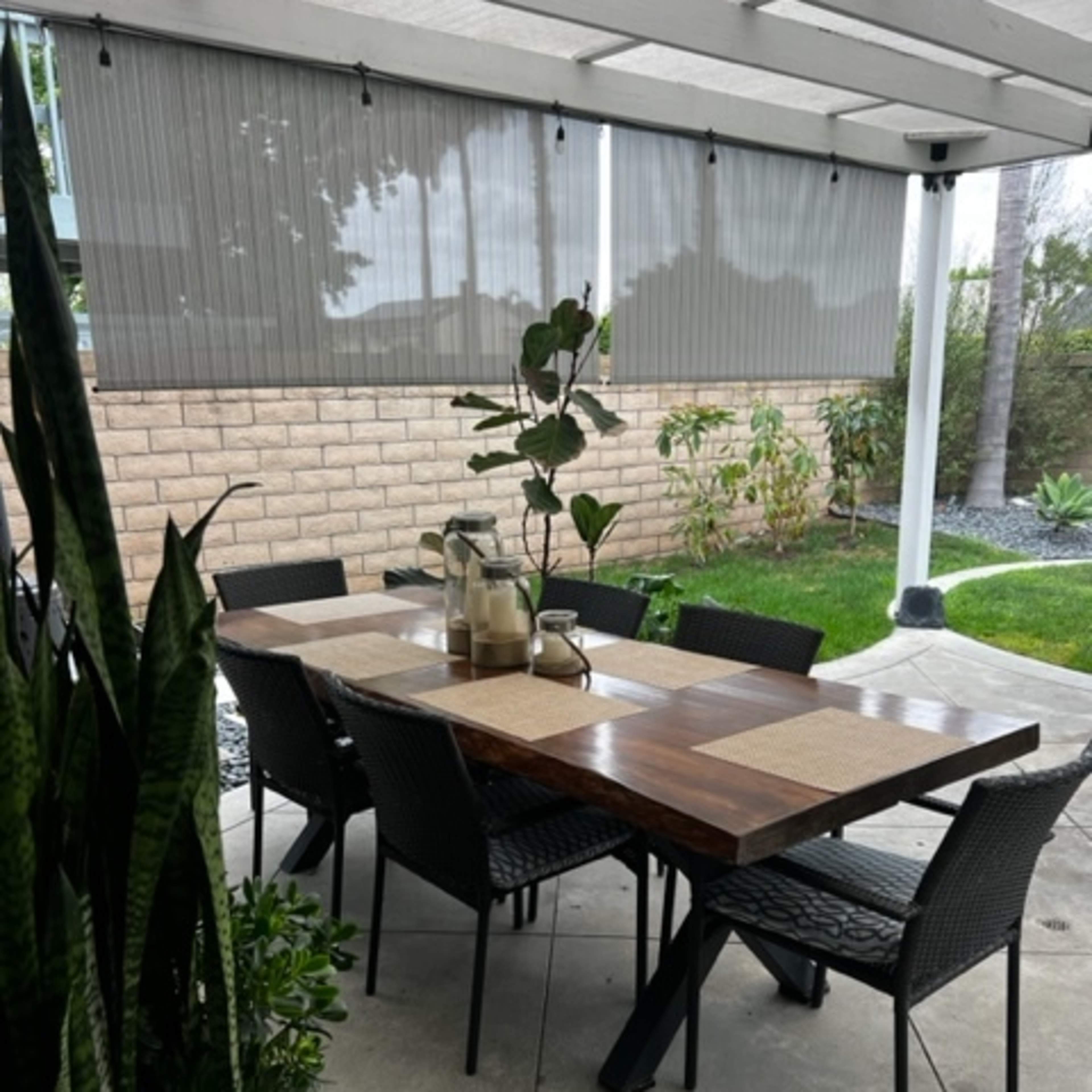 A wooden dining table with woven chairs is set up under a covered patio surrounded by potted plants and a stone wall.