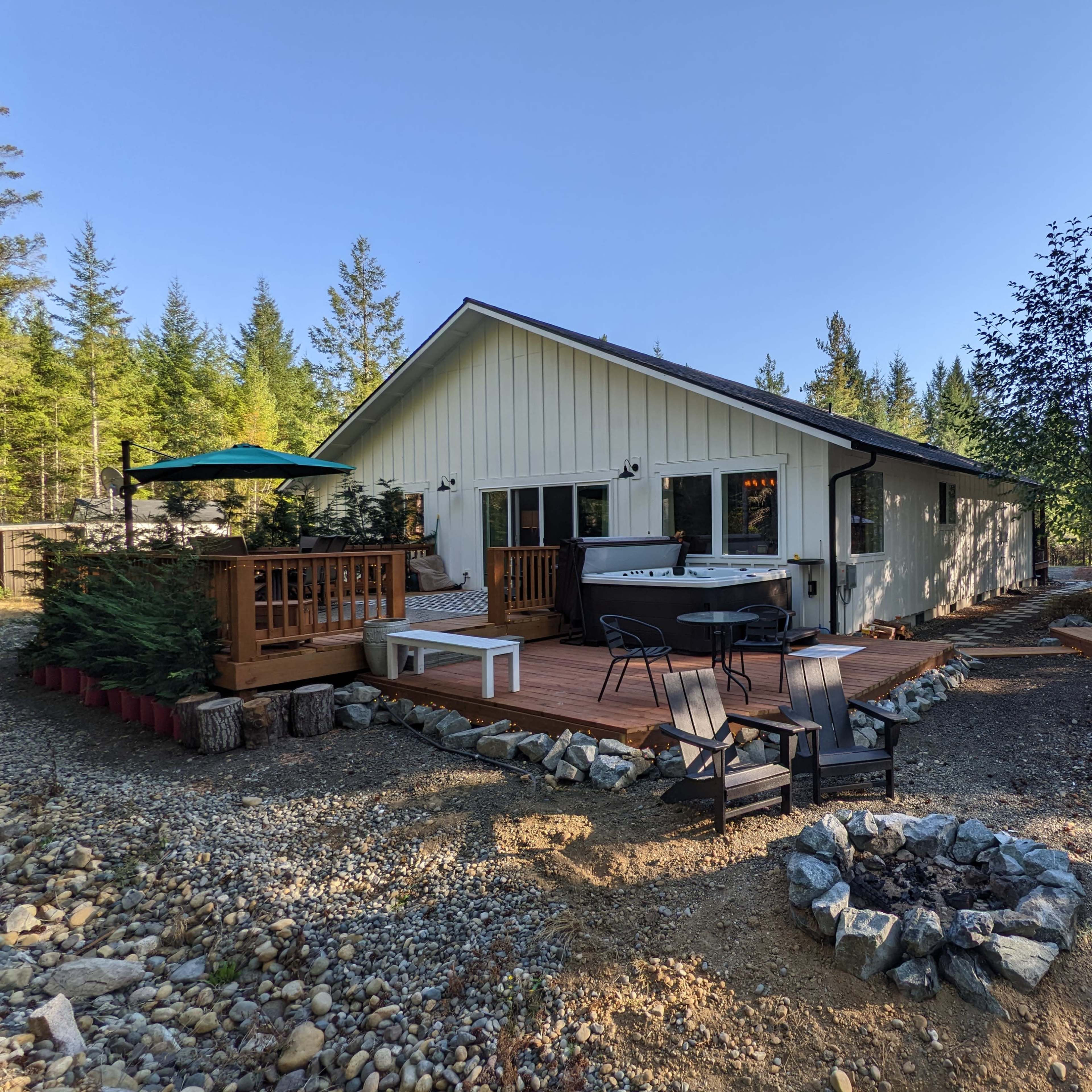 A modern cabin with a wooden deck, outdoor seating, and a hot tub surrounded by trees and a gravel pathway.