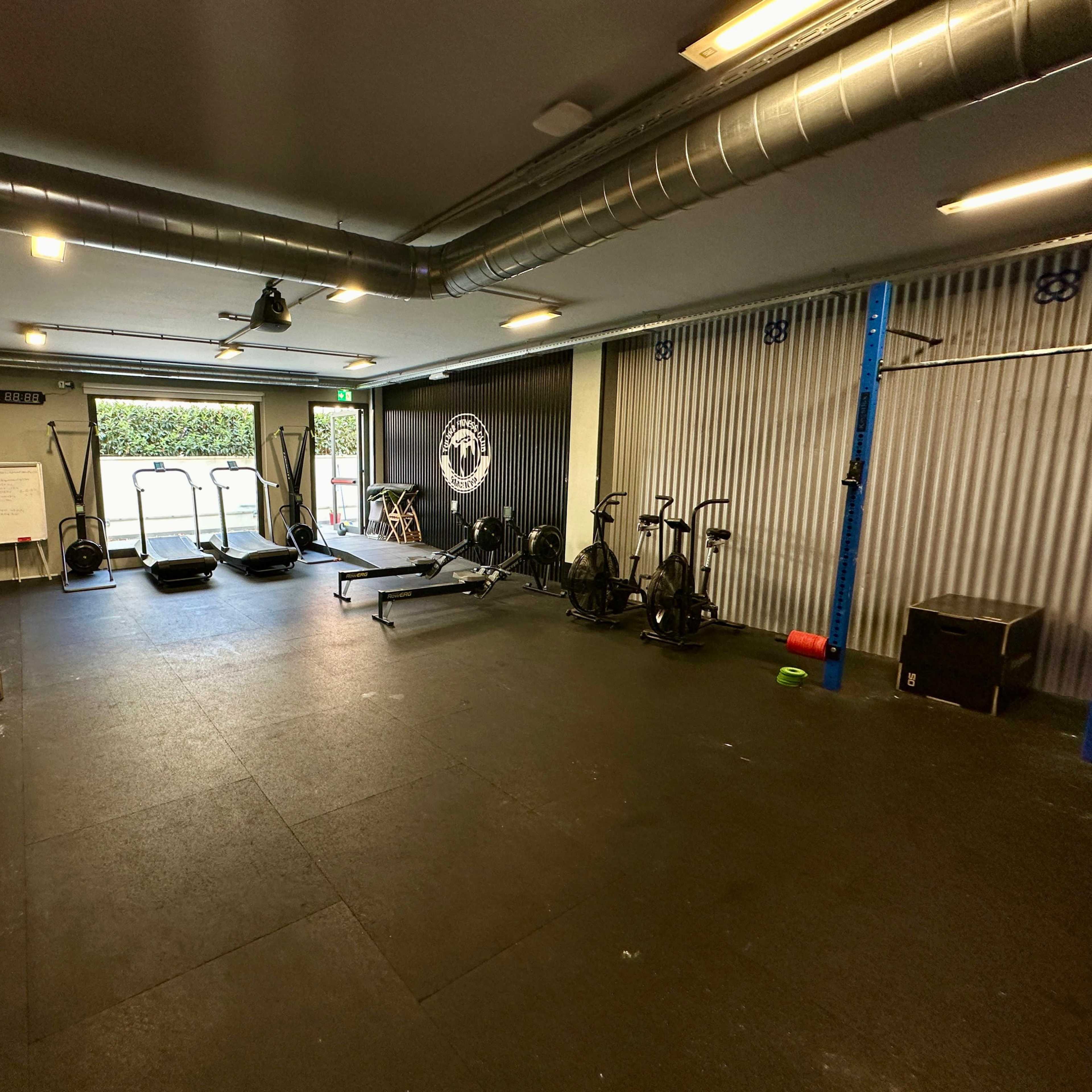 A modern gym with various exercise equipment, including weights, a rowing machine, and sleds, set in an open space with a large window.