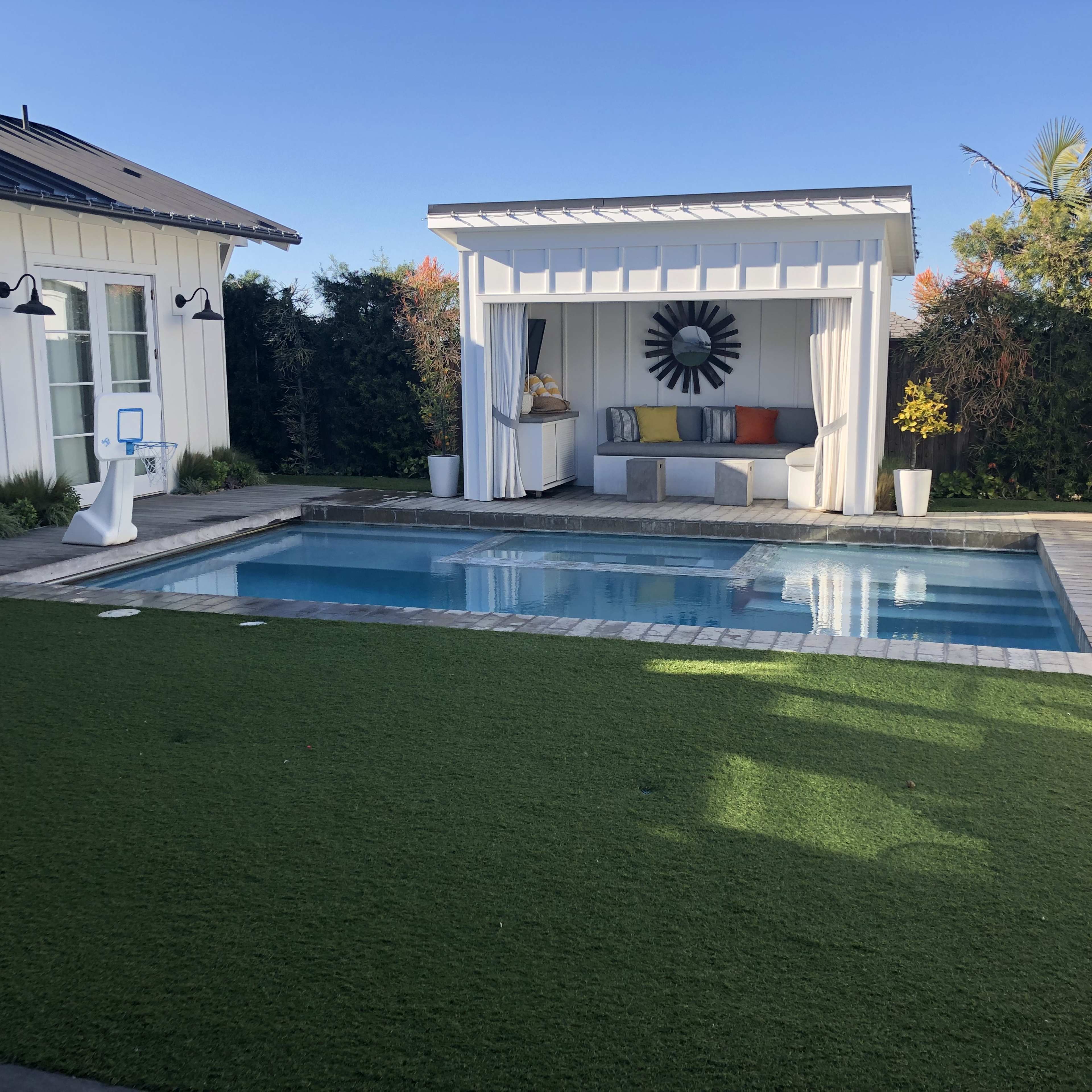 A modern backyard featuring a swimming pool, a decorative outdoor cabana with seating, and a lawn area with artificial turf.
