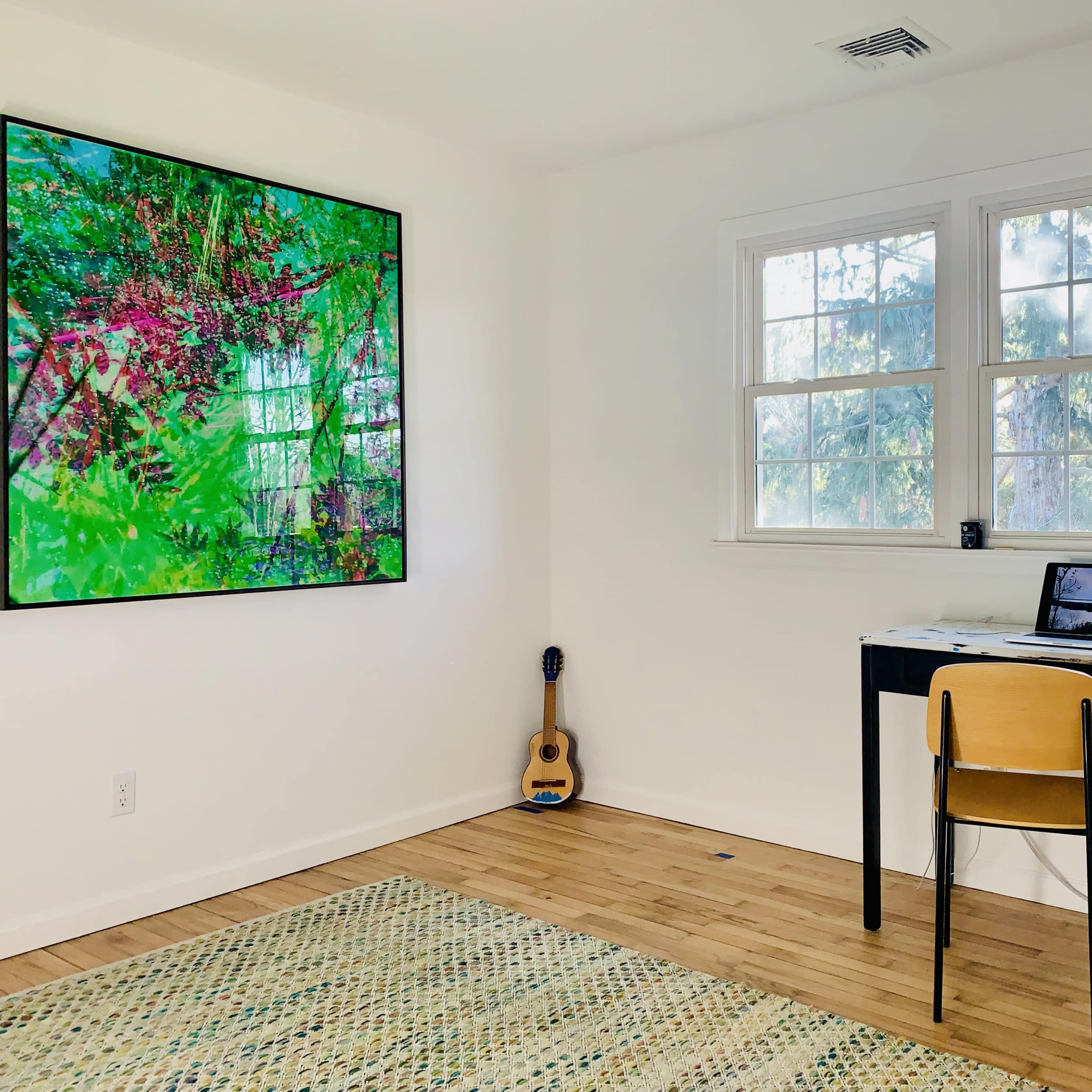 A small room features a desk with a laptop, a chair, a guitar in the corner, and a large colorful artwork on the wall.