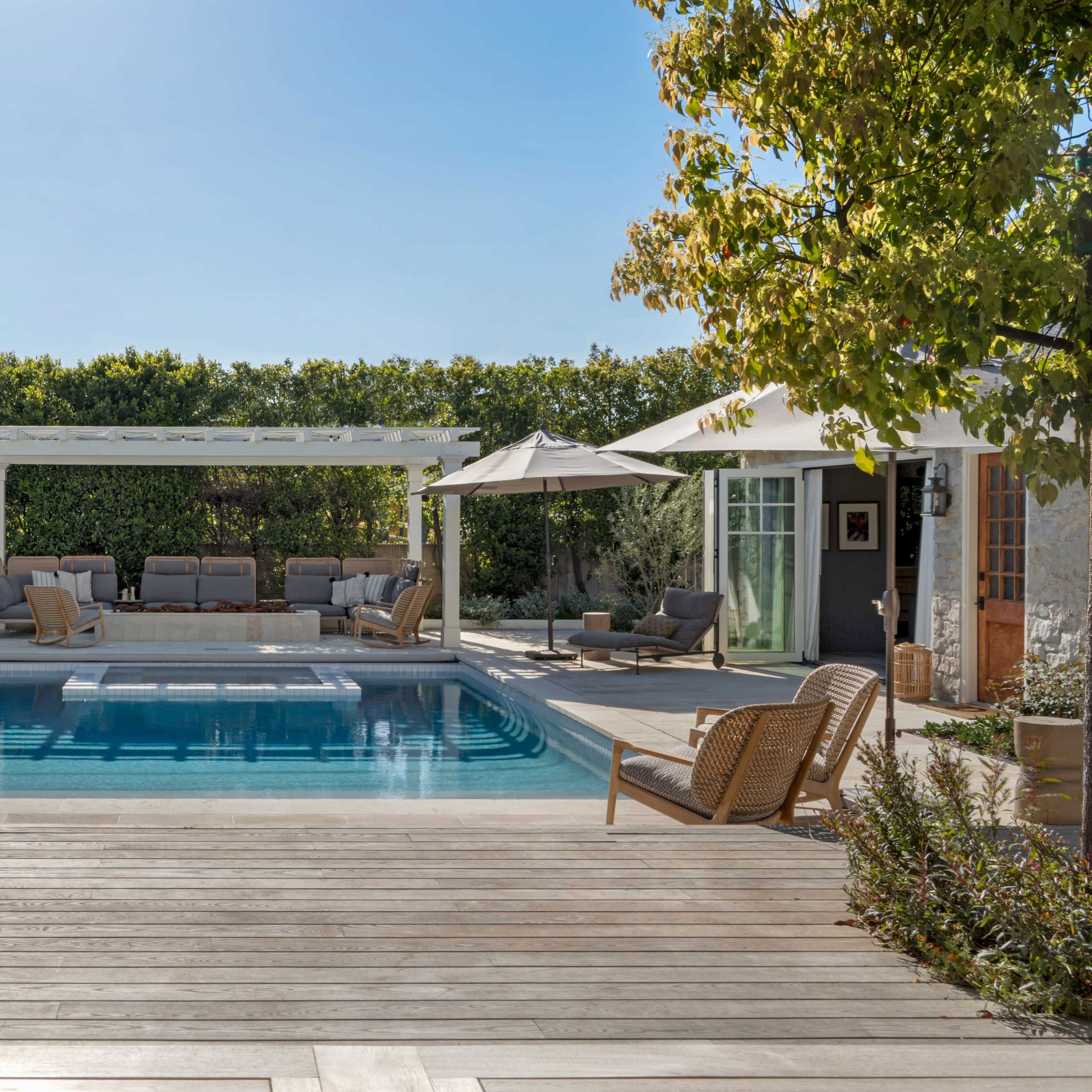 A backyard features a rectangular swimming pool surrounded by seating areas, umbrellas, and lush greenery.