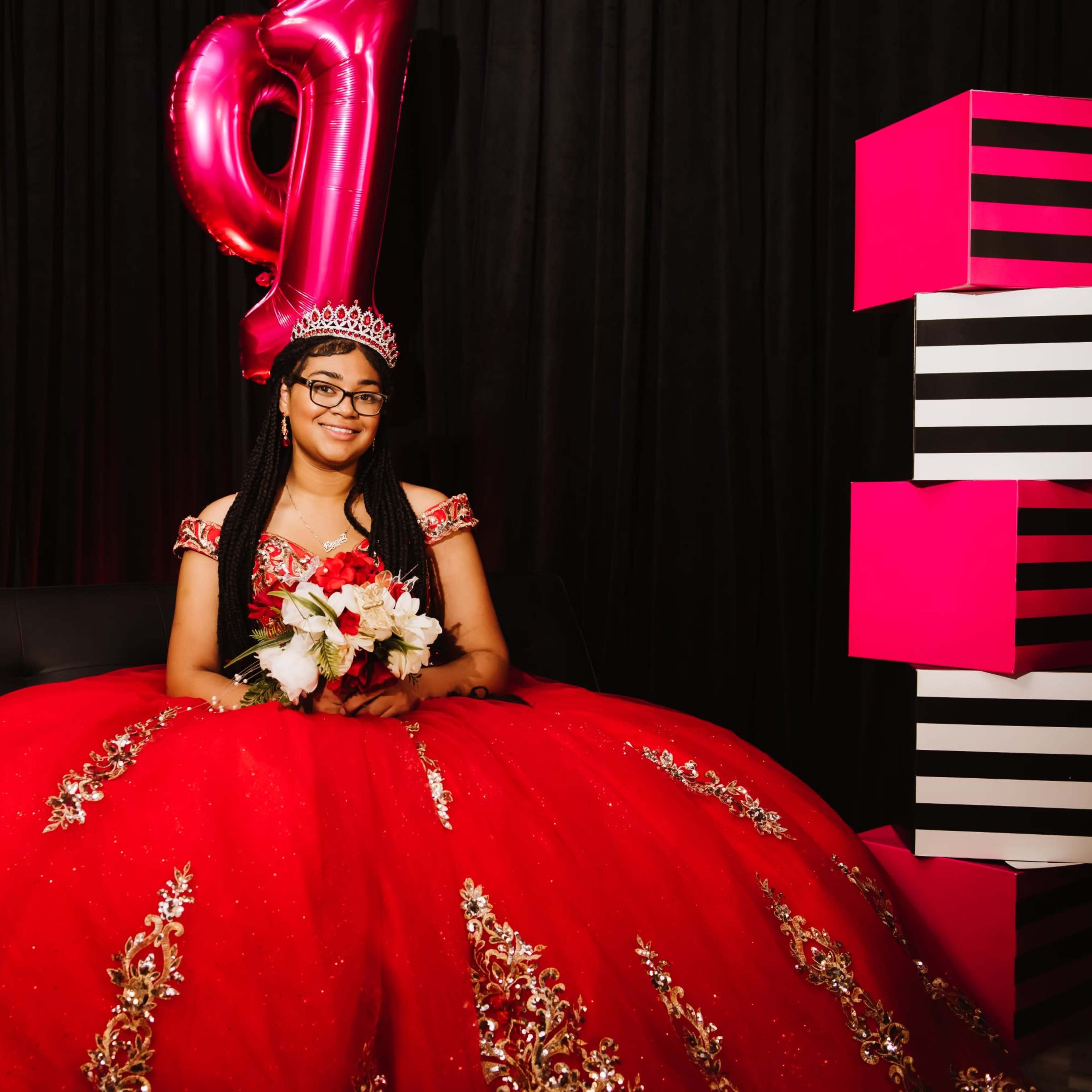 A girl in a red ball gown sits on a couch, holding a bouquet of flowers, with a large pink "16" balloon and black and pink decorations in the background.