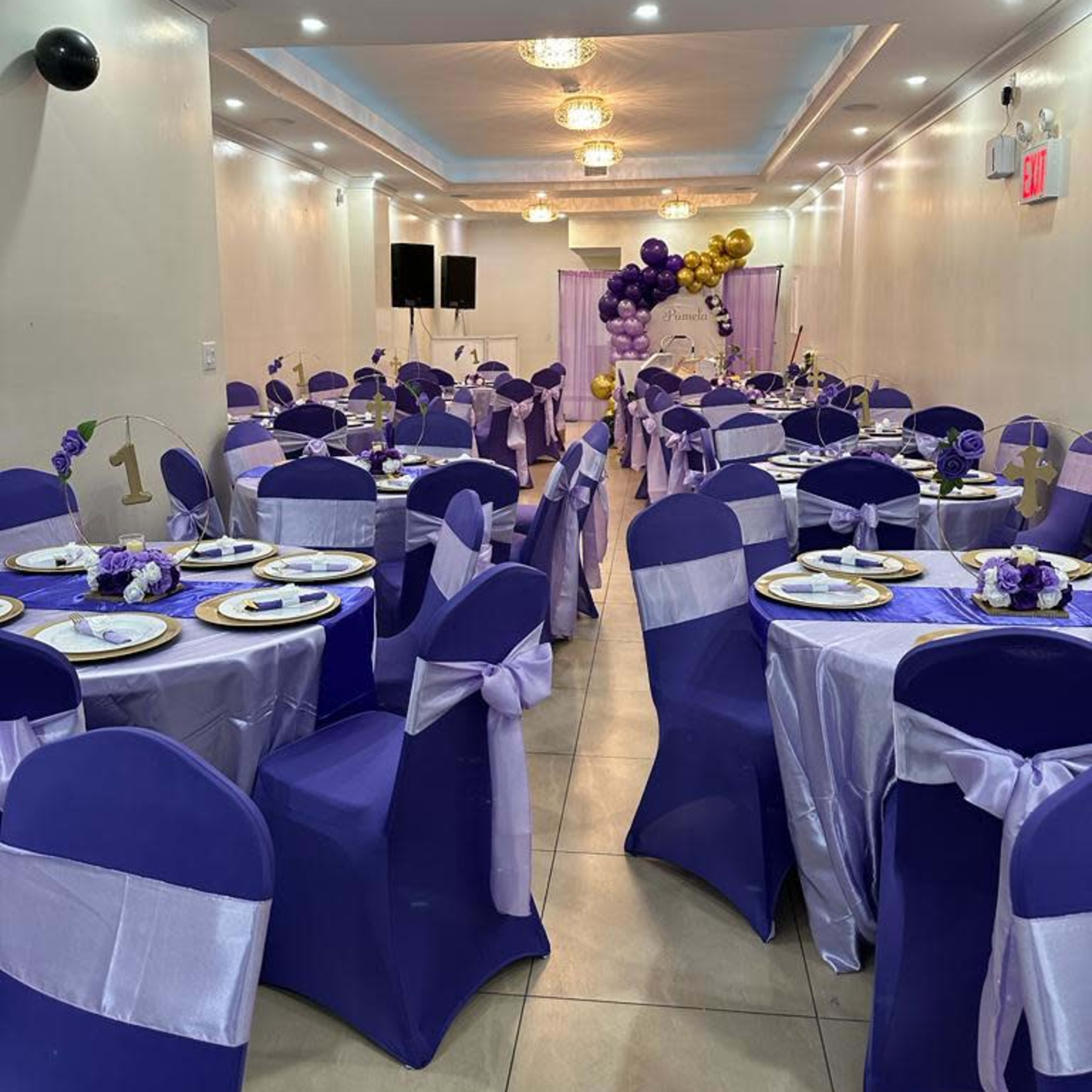 A decorated event space with tables set up in purple and white, featuring balloons and a stage area in the background.