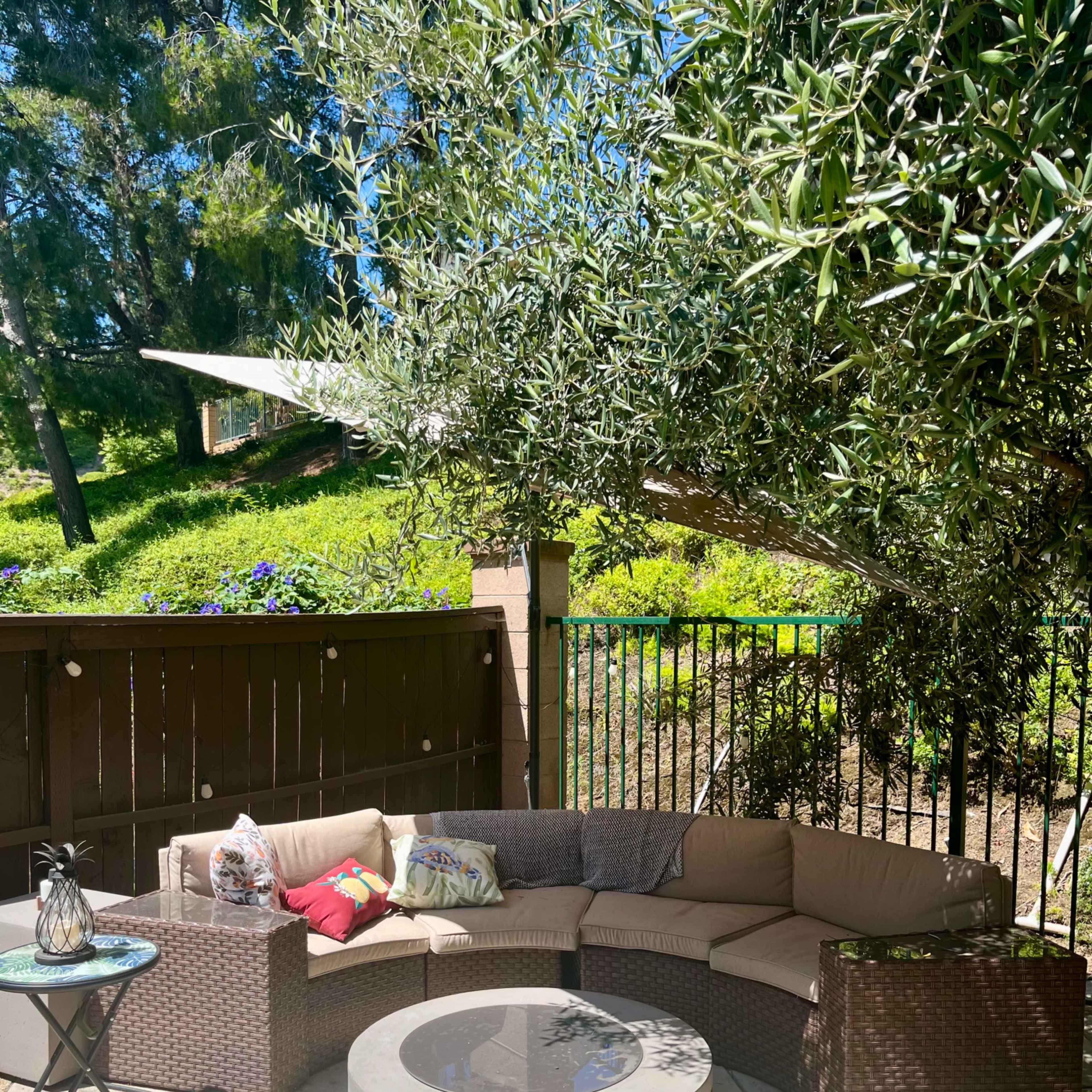 A circular outdoor seating area with a curved sectional sofa, a round coffee table, and a small side table is situated under a tree in a sunny garden.