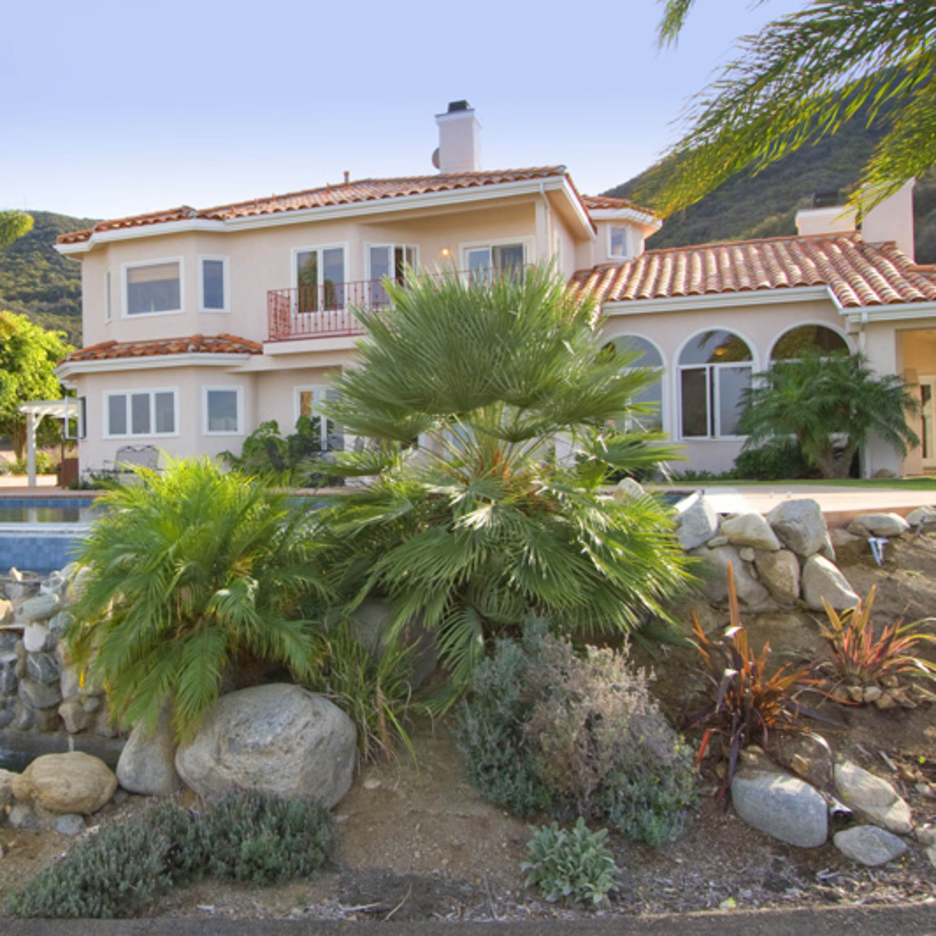 A large house with a red-tiled roof is situated next to a swimming pool and a waterfall, surrounded by tropical plants and mountains in the background.